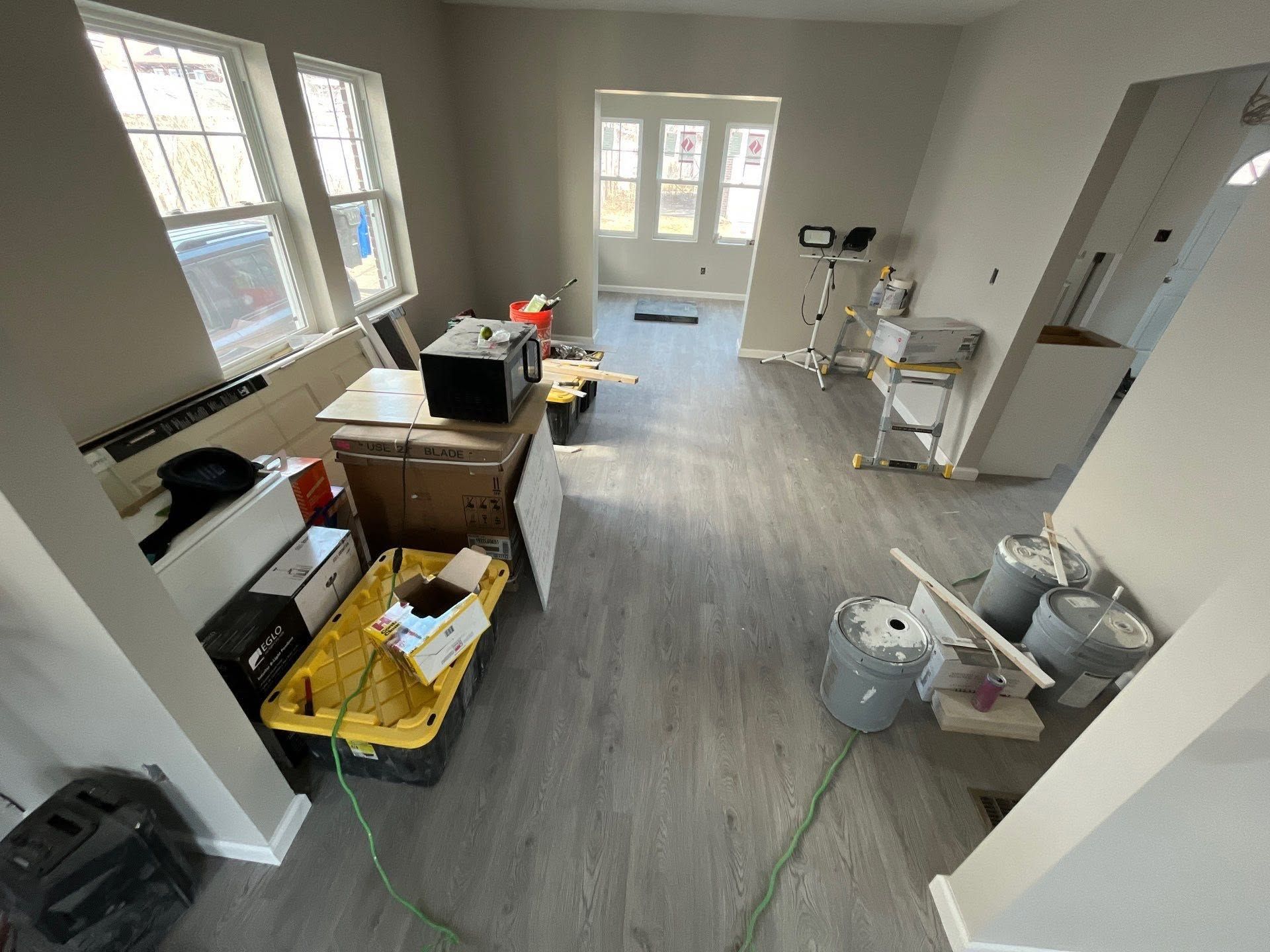 Cluttered room with boxes, tools, and supplies on a gray floor, lit by windows and a back door.
