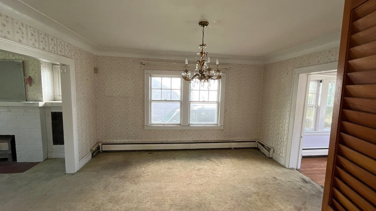 Empty beige dining room with chandelier, carpeted floor, windows, and open doorways.