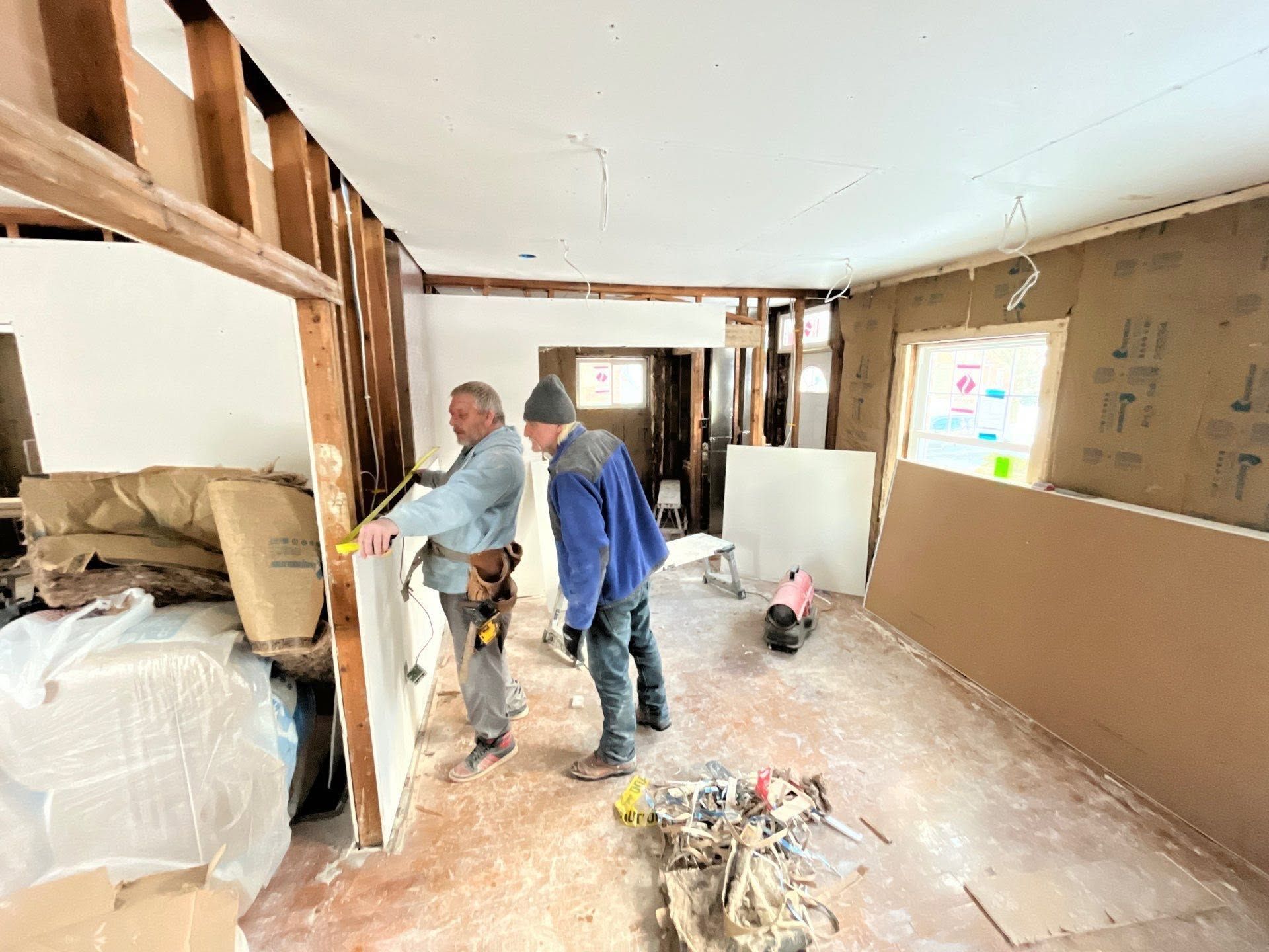 Two workers inspect a gutted room under renovation, with debris, exposed framing, and unfinished walls.