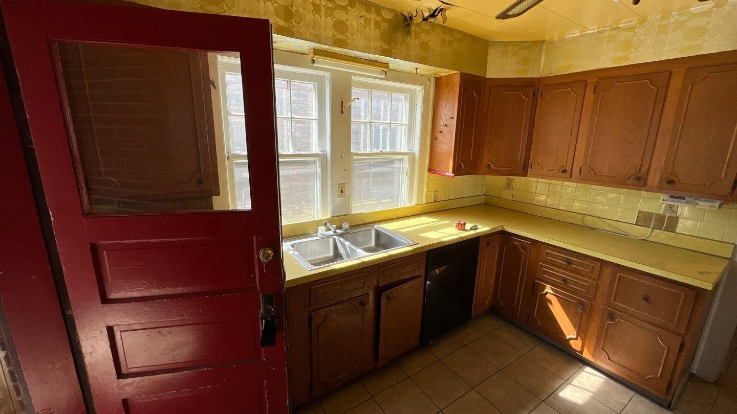 Small kitchen with red door, wooden cabinets, white sink under a window, and worn yellow walls.
