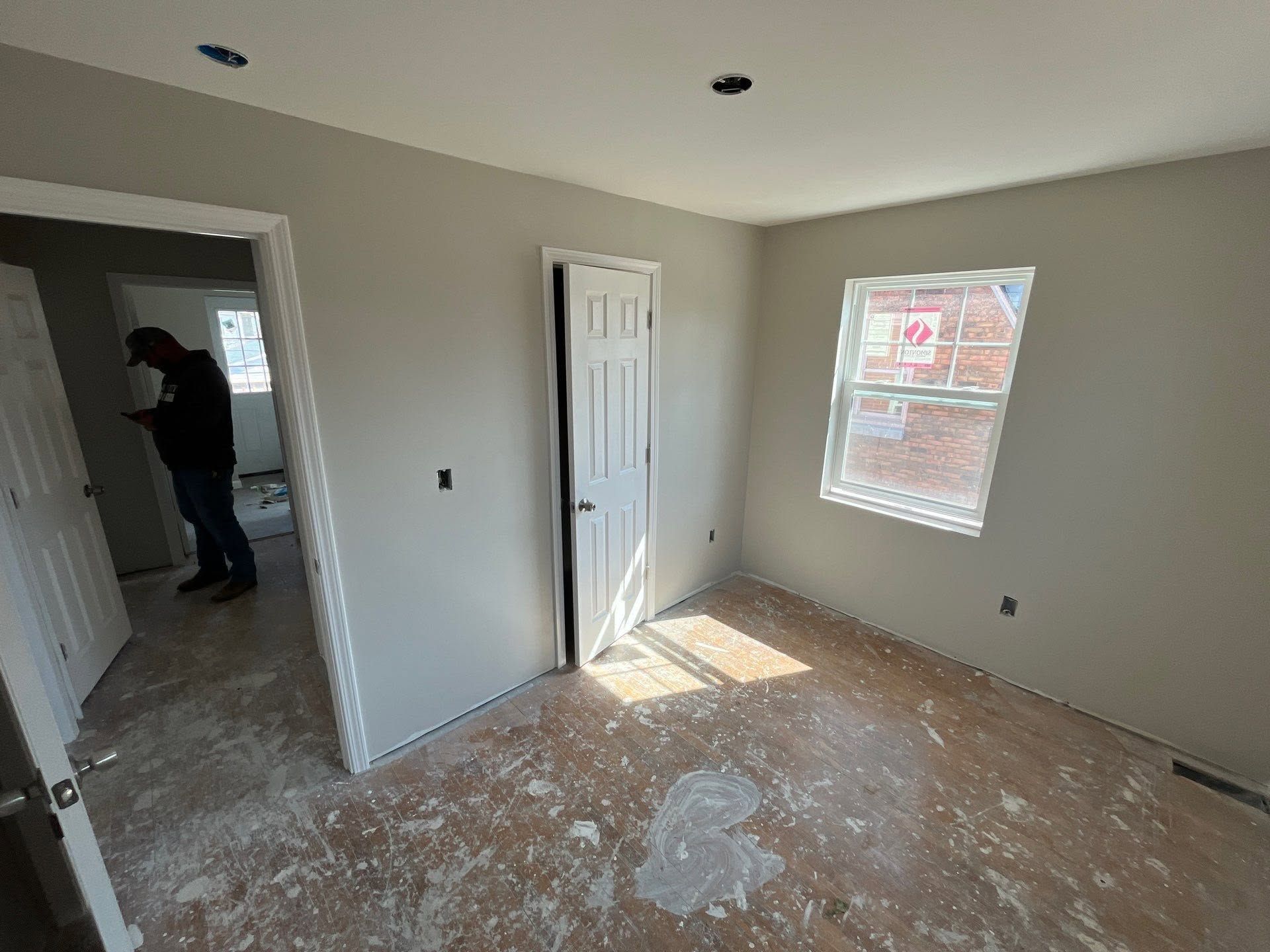 Empty room under renovation with unfinished drywall, window light, white door, and person visible in hallway mirror
