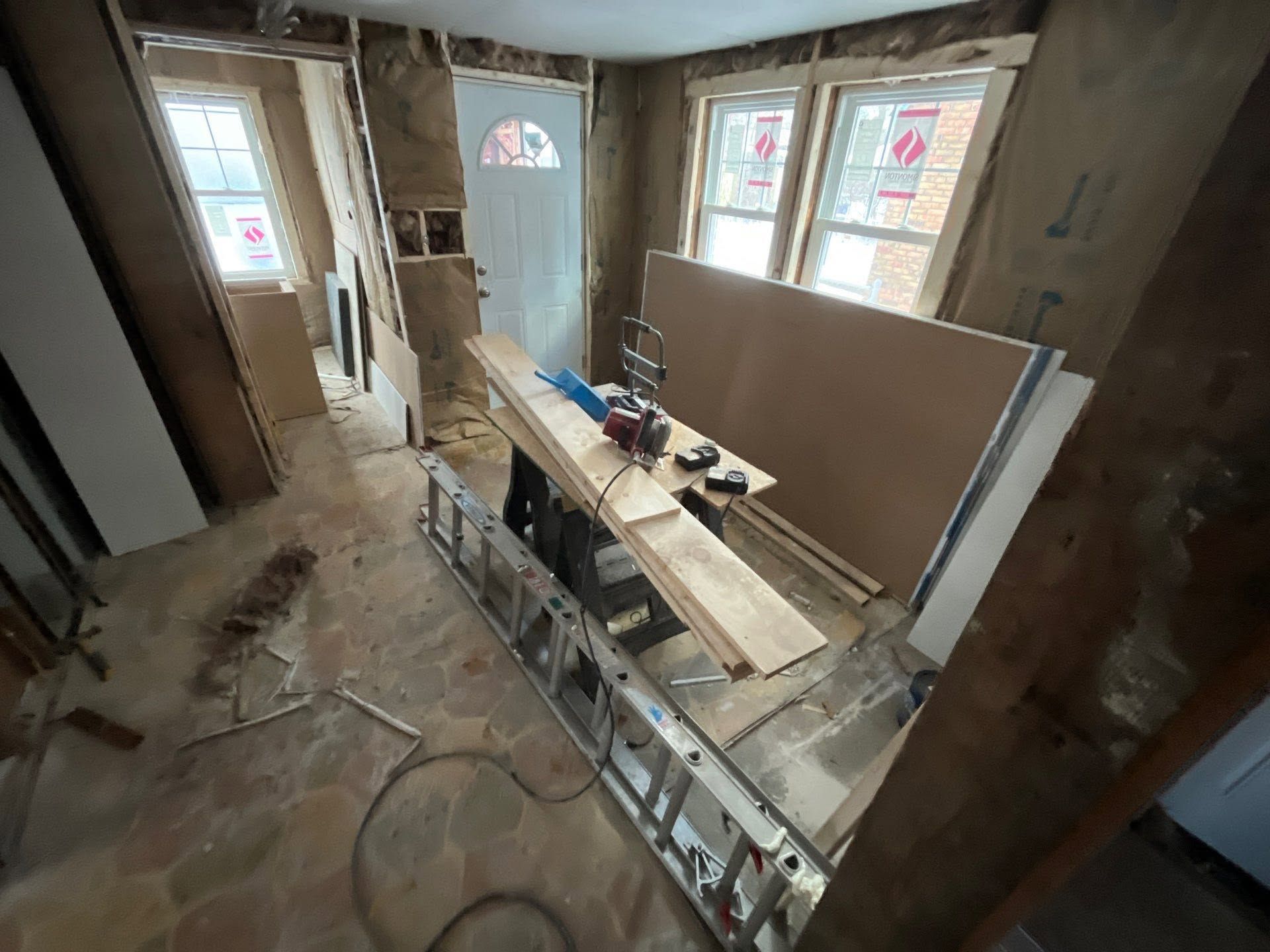 Partially renovated room with drywall, tools, and a workbench near two windows and a front door.