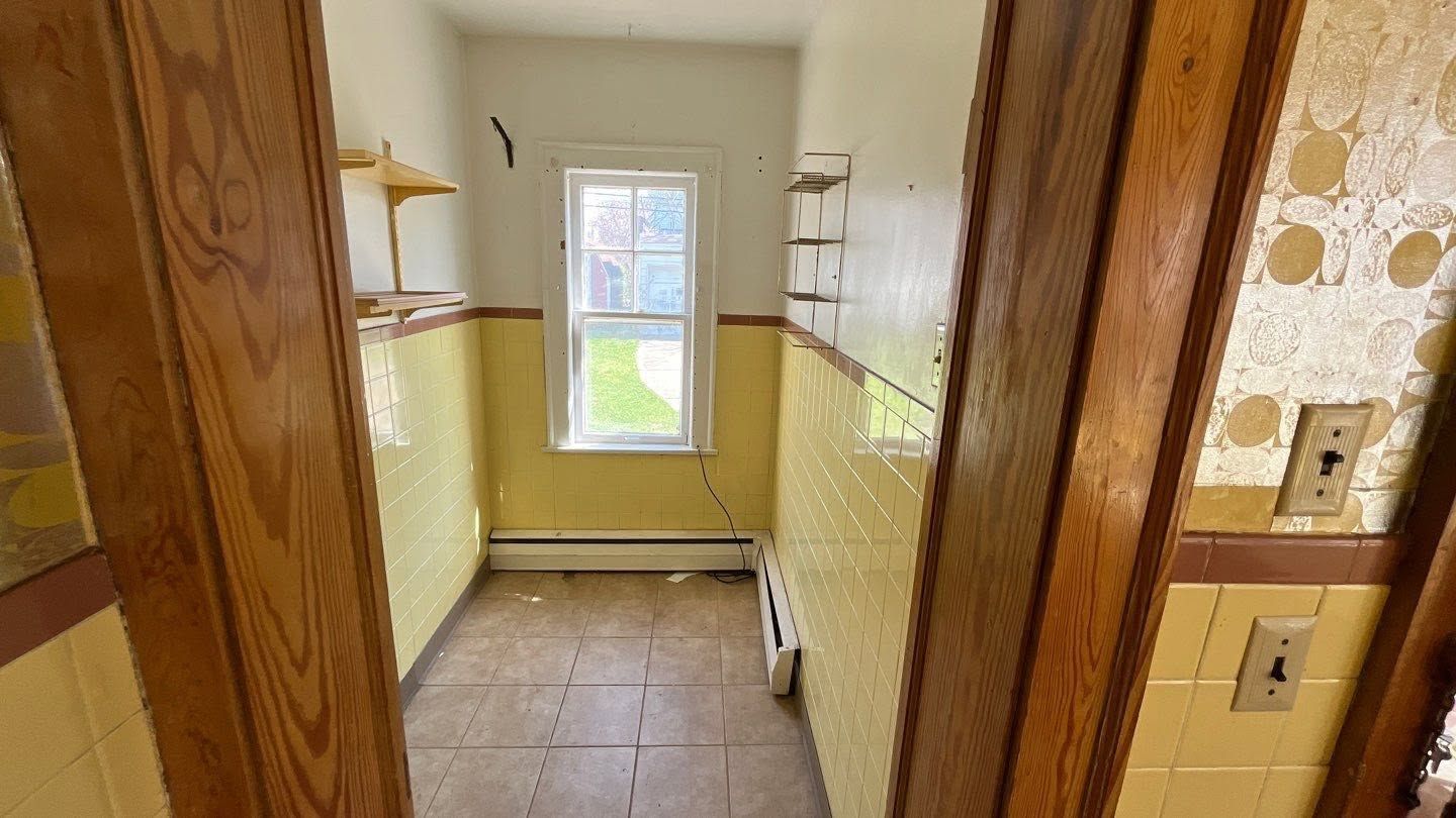 Narrow empty kitchen with tiled walls, window at the end, and wooden door frames.