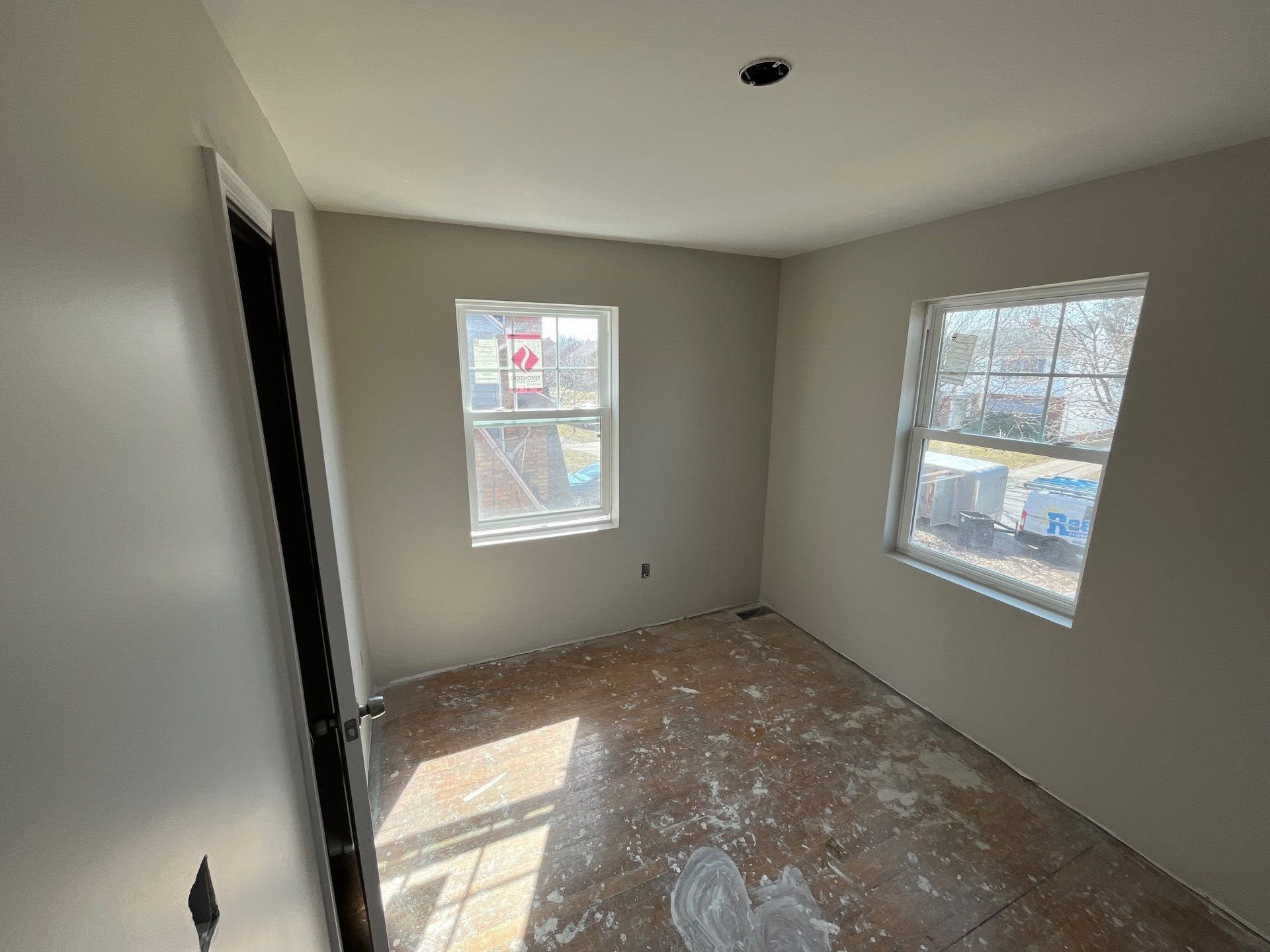 Empty room with gray walls, two windows, and a paint-splattered floor in sunlight