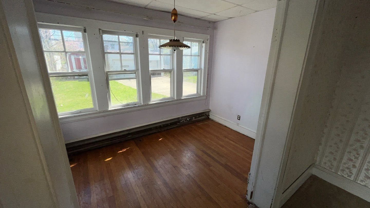 Empty sunlit room with hardwood floors, large windows, and white walls