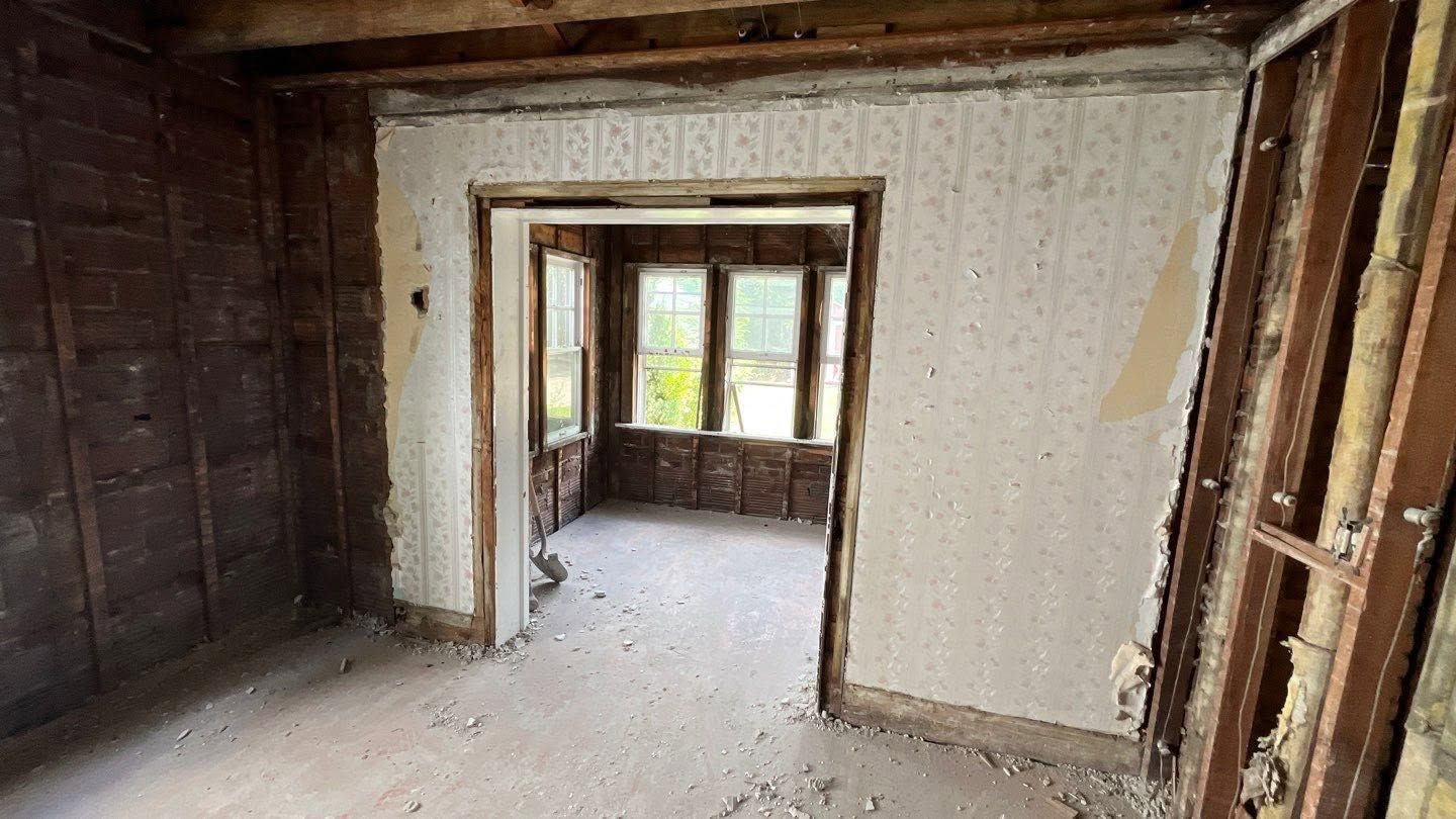 Empty, dilapidated room with exposed wood, white walls, debris on the floor, and a doorway to a sunlit bay window area