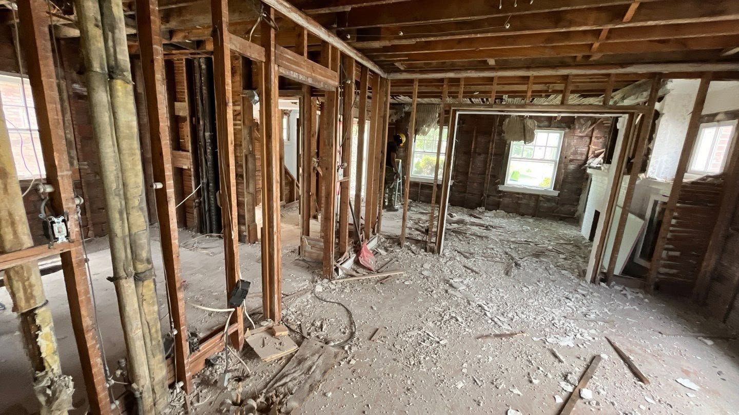 Interior of a gutted house under renovation, with exposed studs, debris-covered floor, and daylight through windows