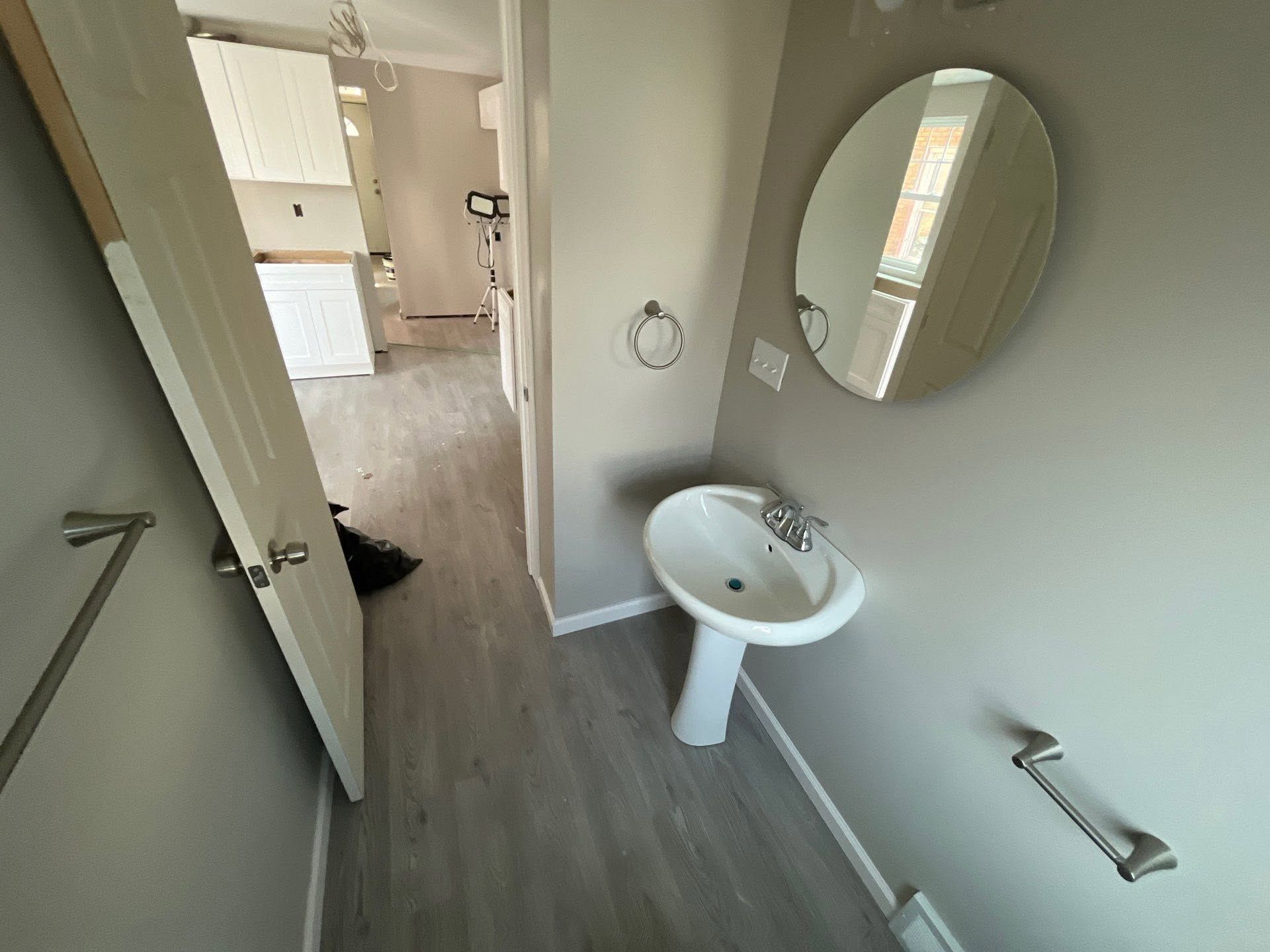 Small bathroom with a pedestal sink, round mirror, and gray floor, viewed toward a hallway.