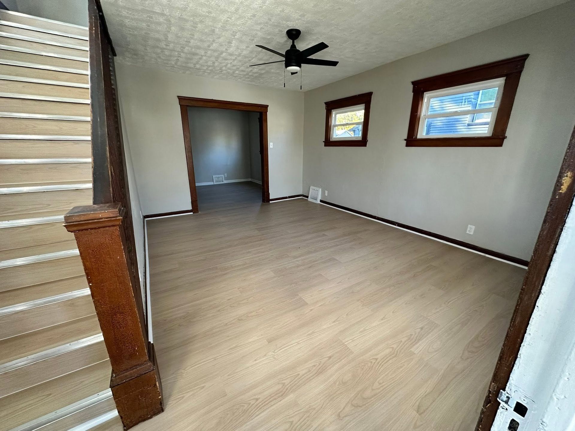 Empty living room with light wood floors, two windows, ceiling fan, and staircase at left.