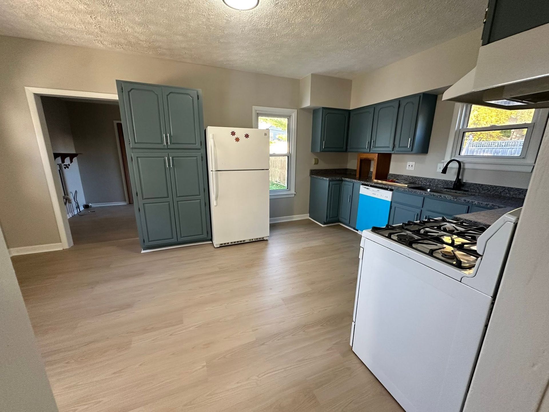 Empty kitchen with light wood floor, teal cabinets, white fridge, and gas stove by a window.
