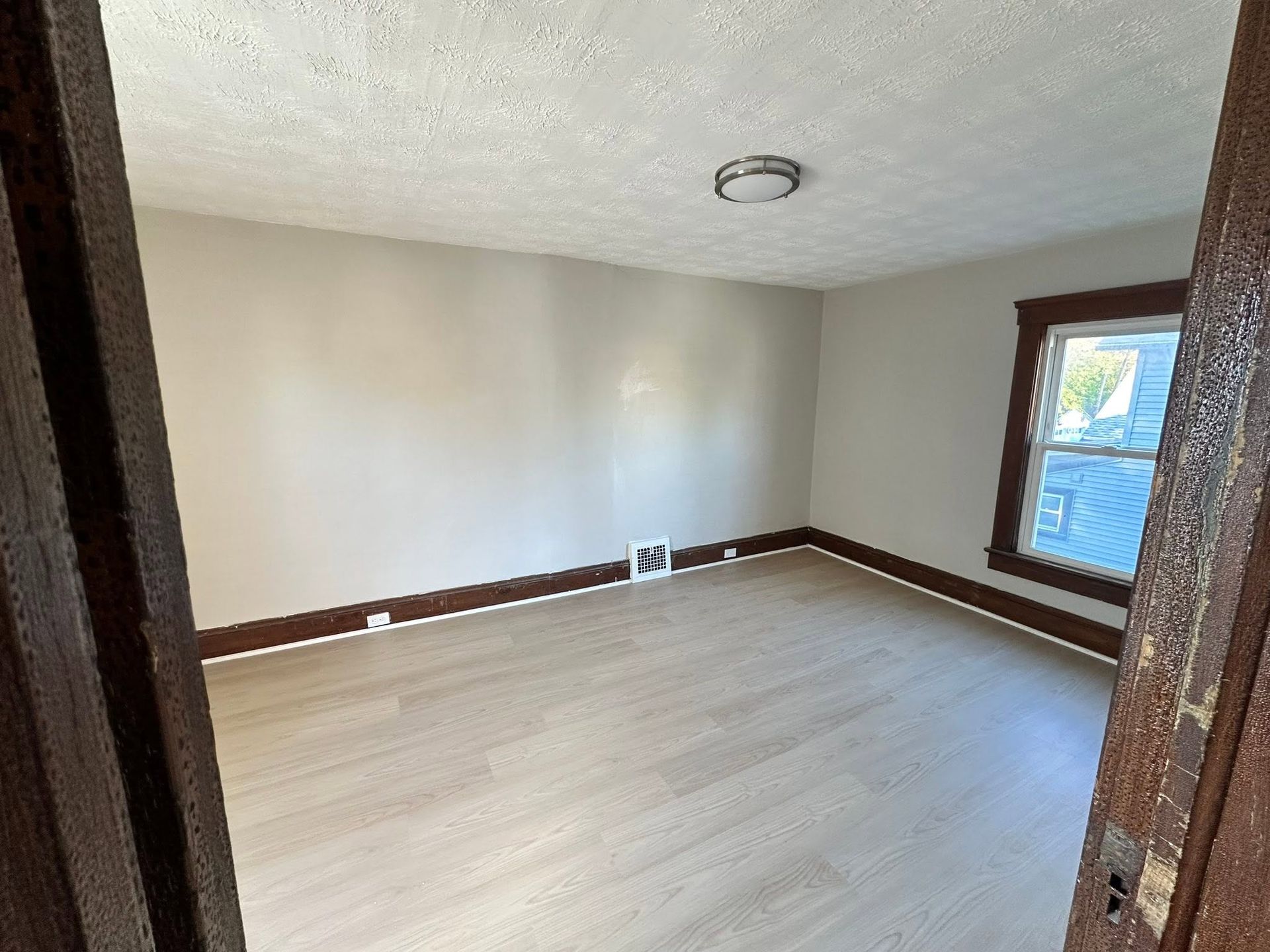 Empty beige room with light wood floor, brown trim, and a window on the right.