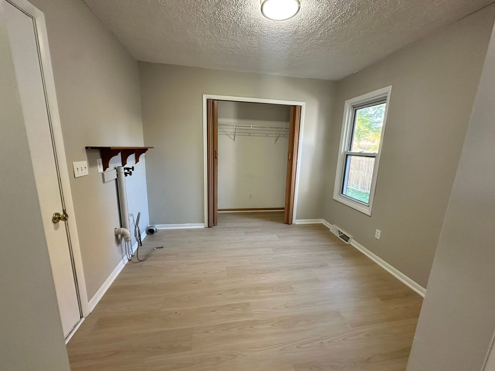 Empty bedroom with beige walls, light wood floor, closet, window, and small wall shelf with hooks.