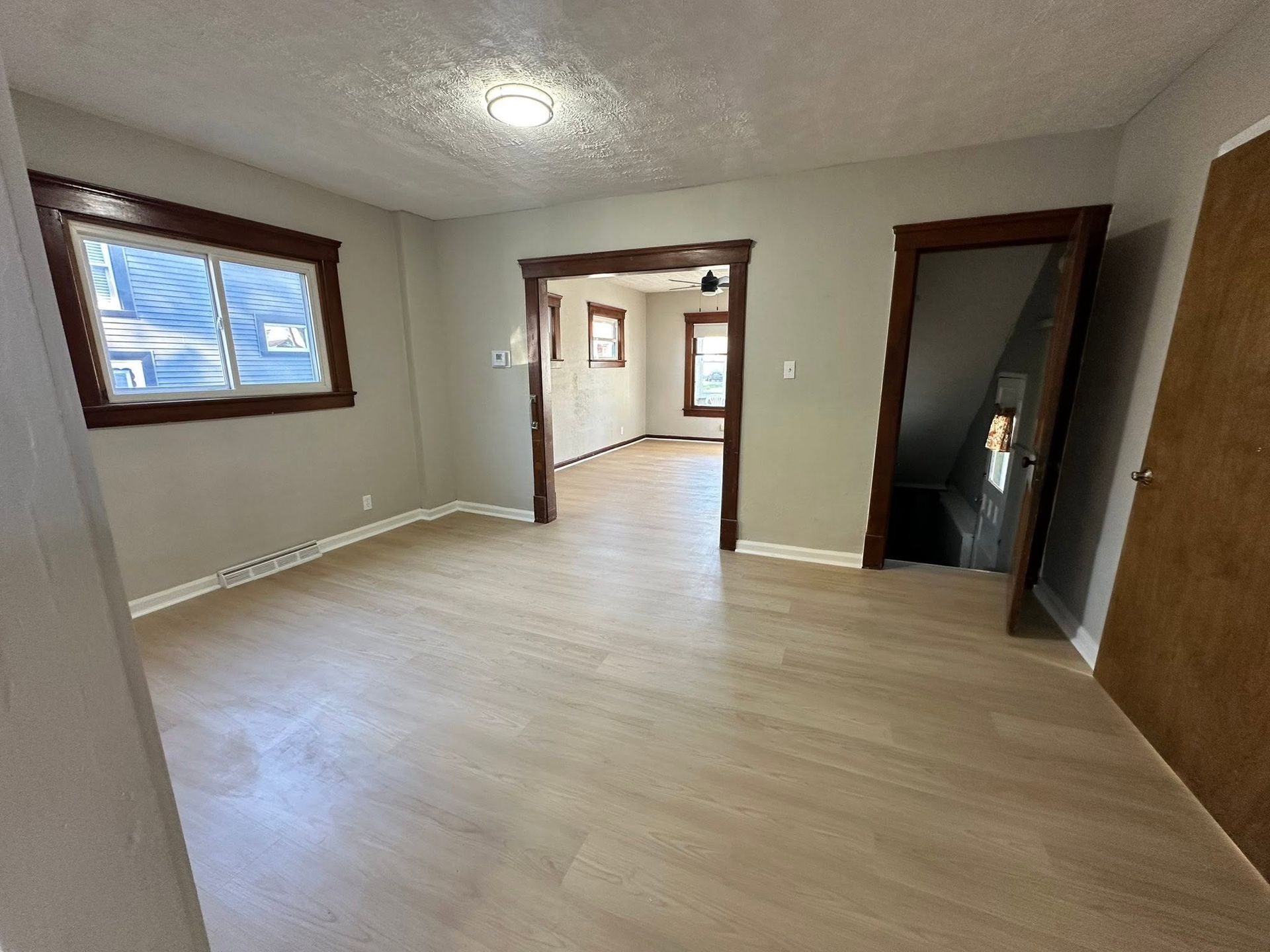 Empty room with light wood floors, beige walls, wood-trimmed windows, and an open doorway to another room