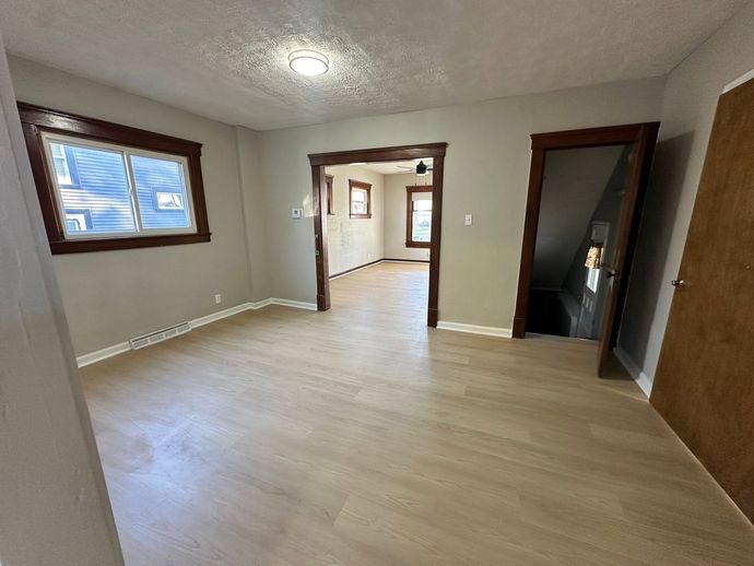 Empty beige room with wood trim, light flooring, and windows, viewed toward an open doorway and hall.