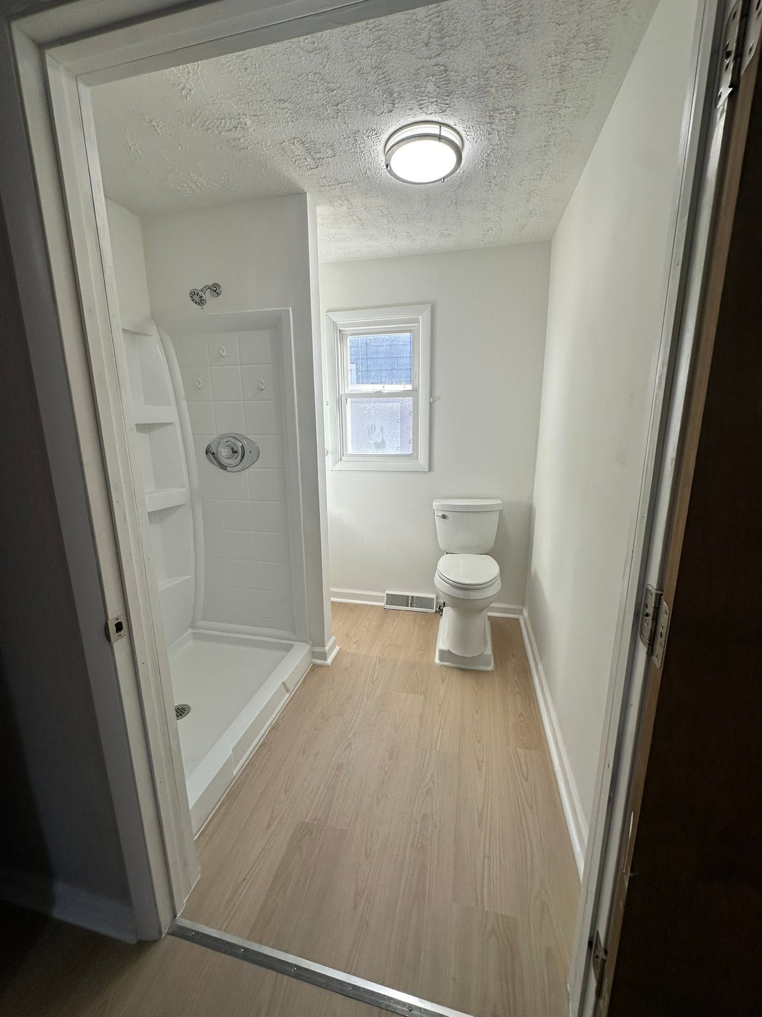 Small bathroom with shower, toilet, window, and wood-look floor tiles.