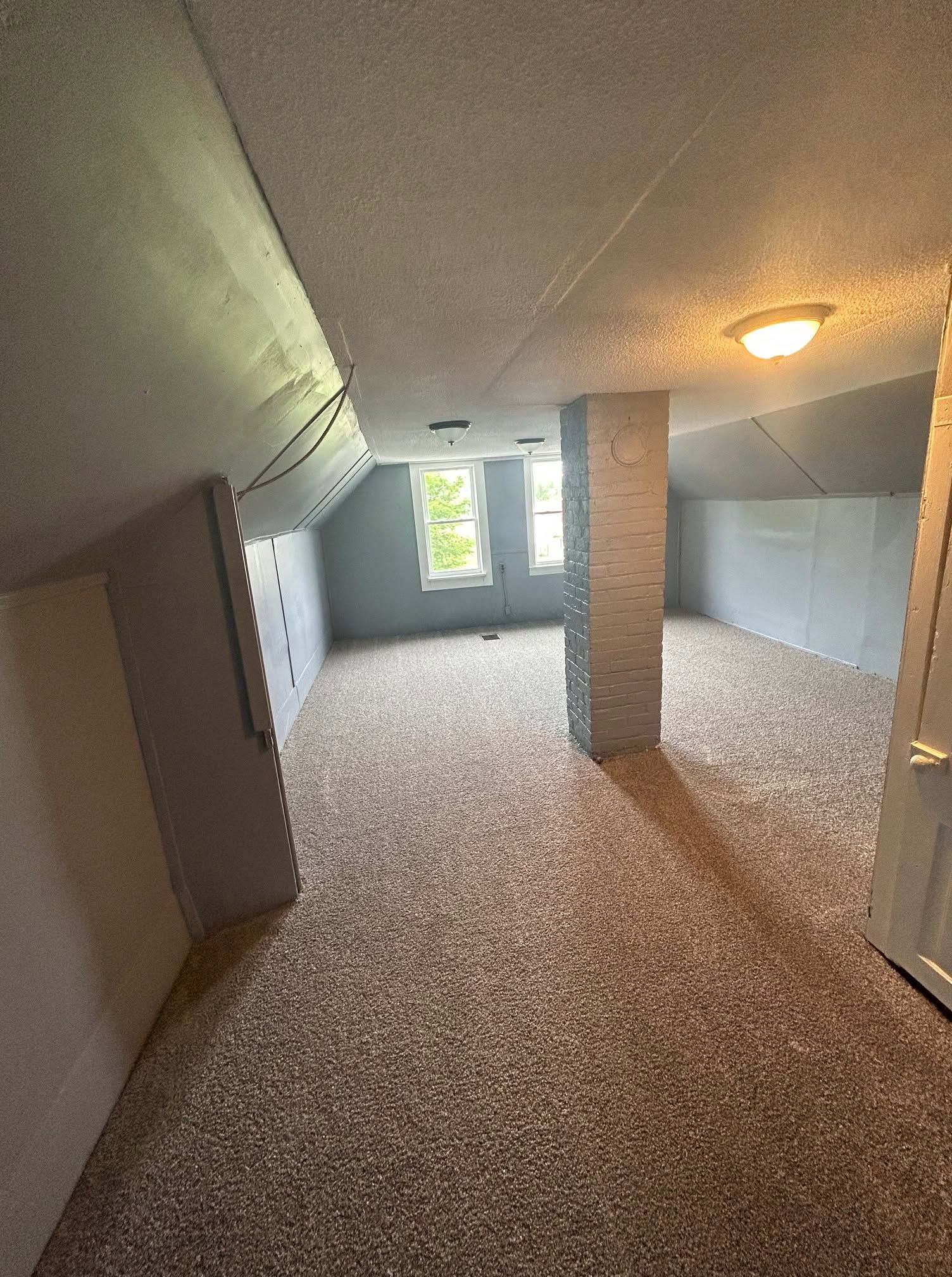 Empty attic room with sloped ceiling, carpeted floor, central support column, and window at the far end.