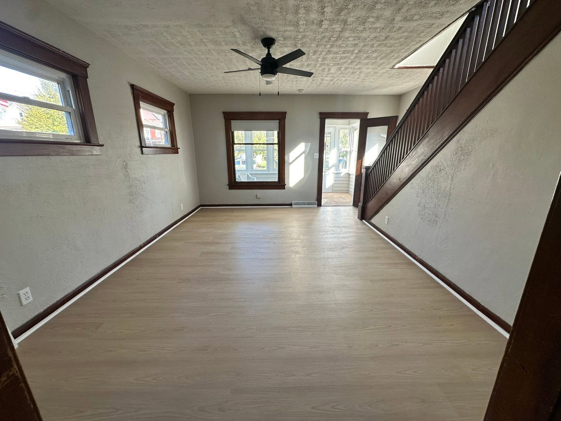 Empty living room with wood flooring, white walls, and ceiling fan, viewed toward windows and a glass door