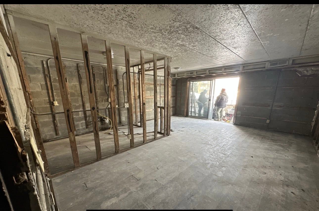 Unfinished basement with exposed wall studs and concrete floor; two people stand near the bright doorway.