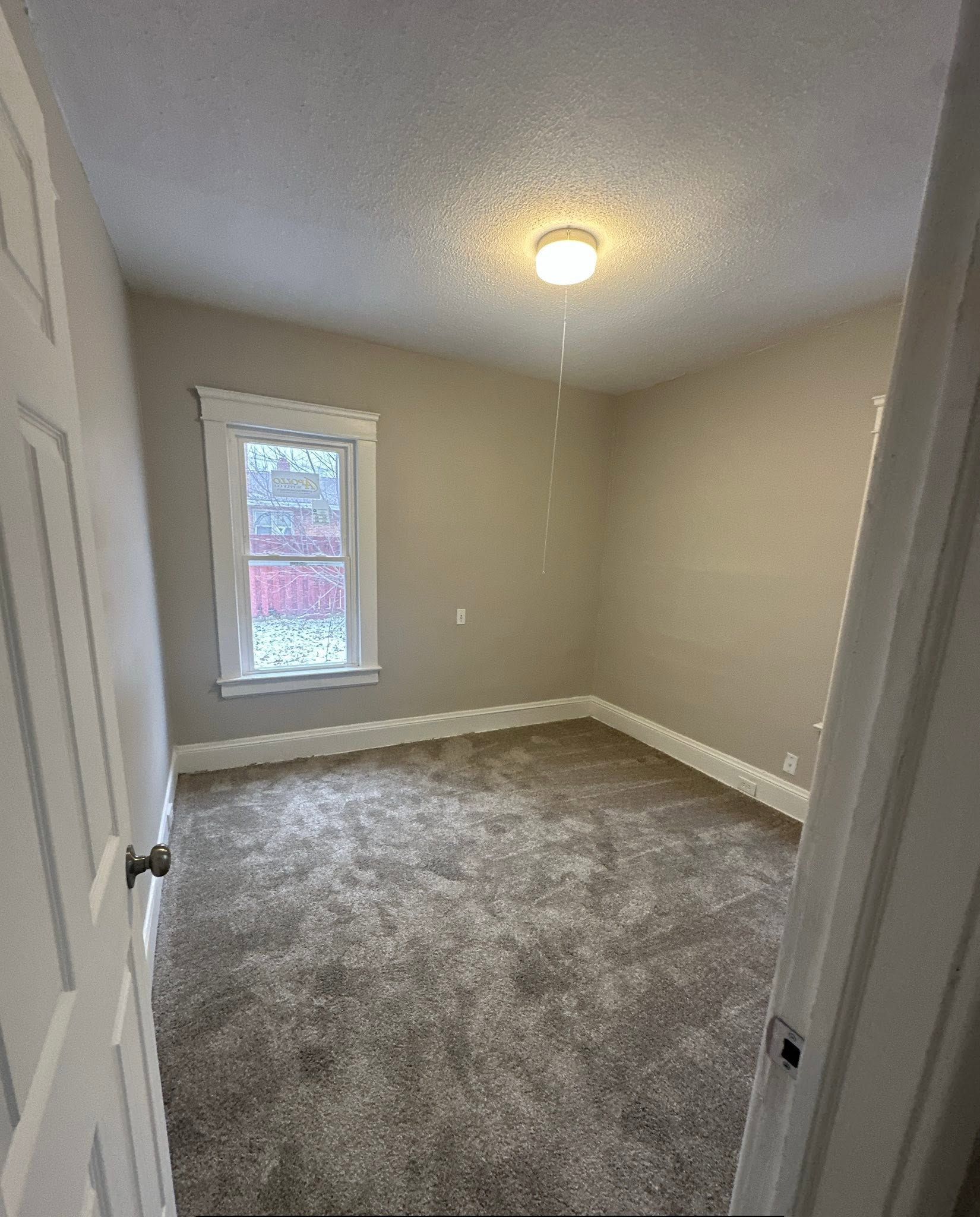Empty beige bedroom with gray carpet, a small window, and a ceiling light on.
