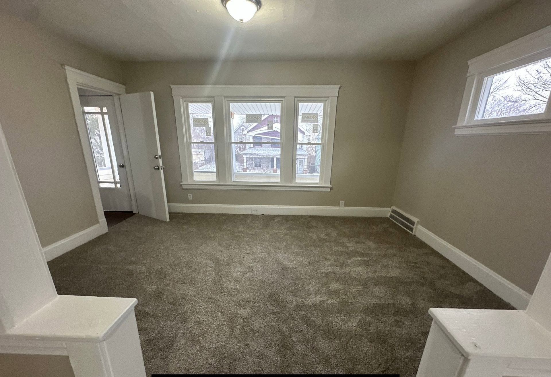 Empty carpeted living room with large front windows and a small side window, viewed from the doorway