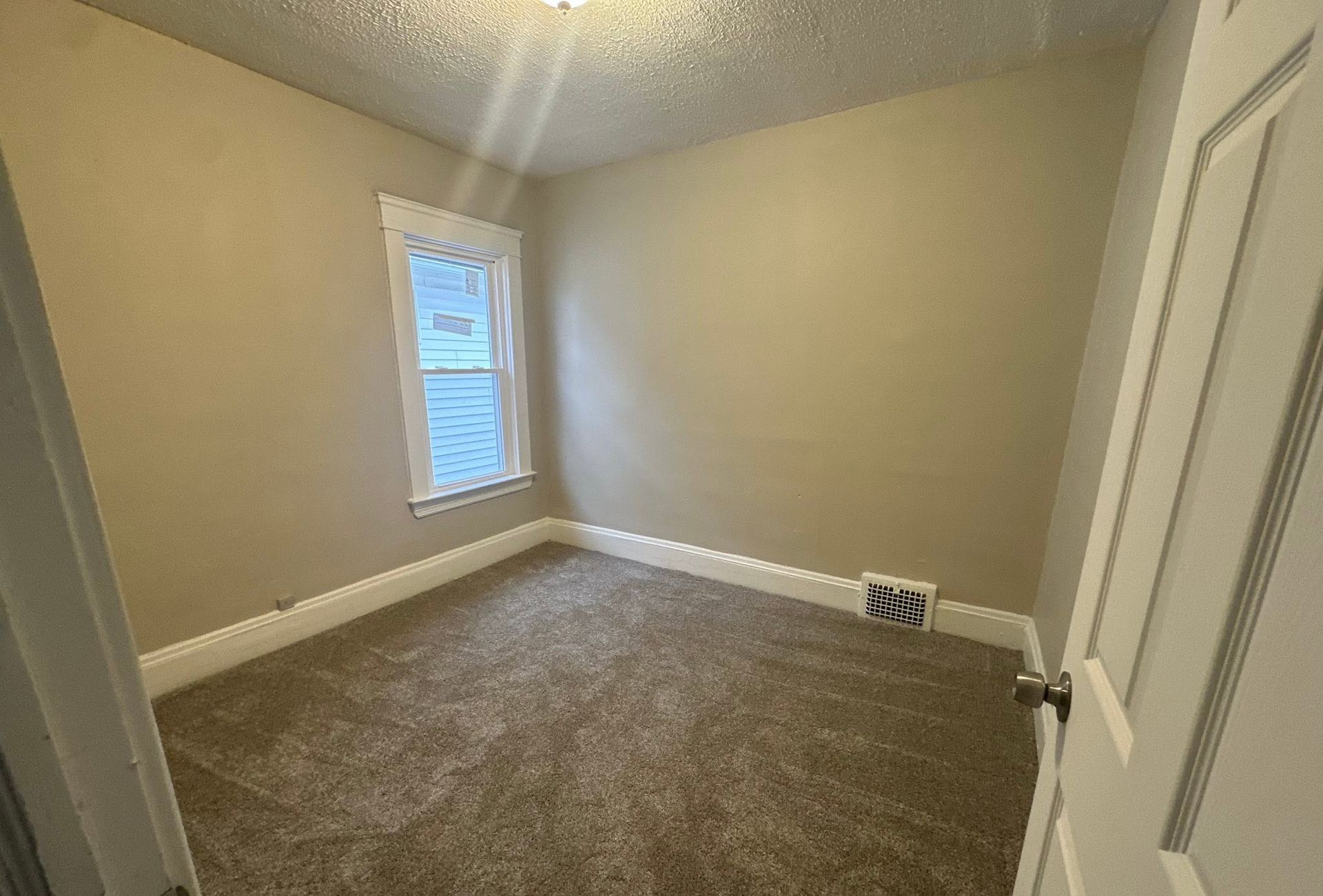 Empty beige carpeted bedroom with a small window and white closet doors.