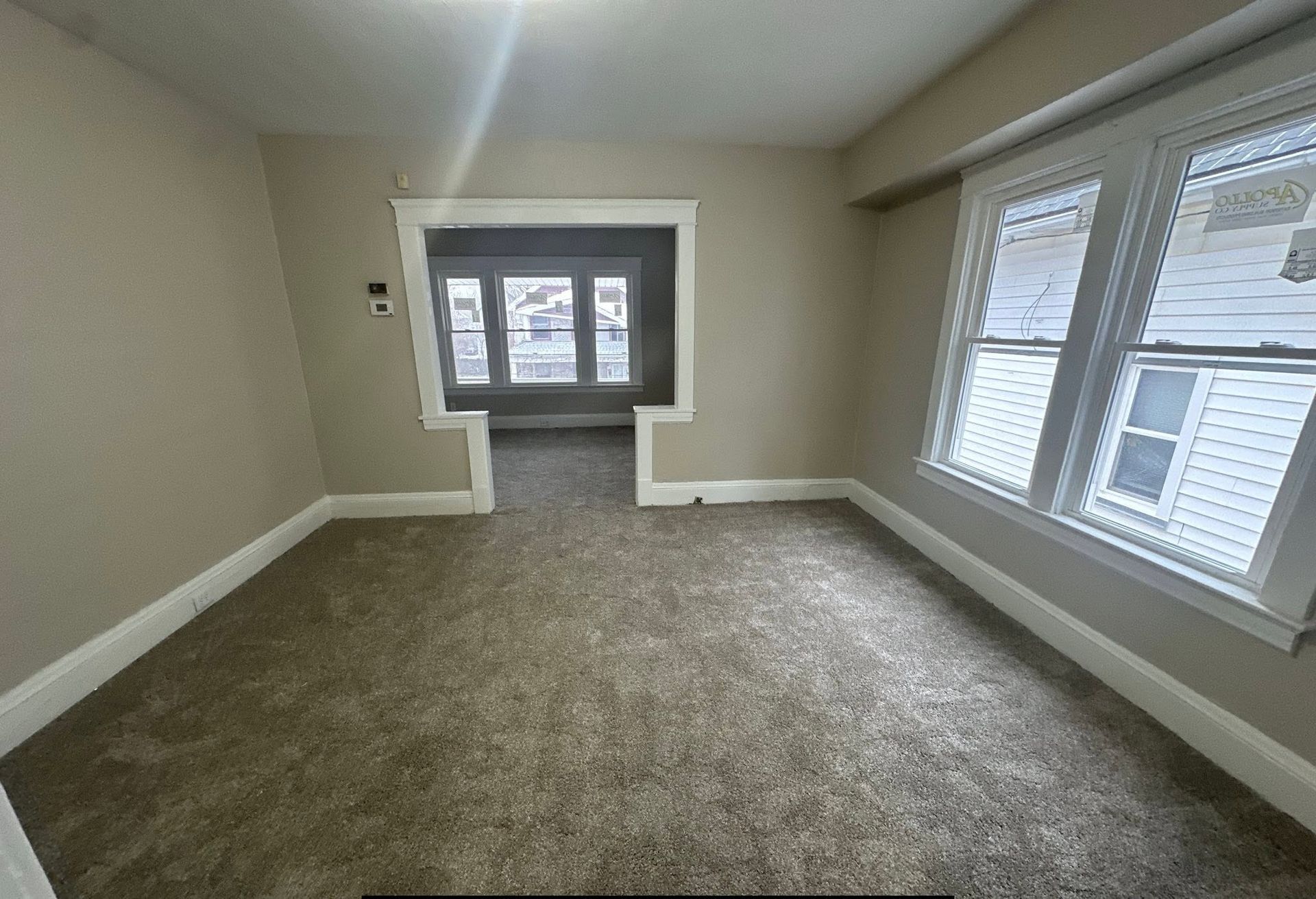 Empty beige carpeted room with white-trimmed windows and a small doorway at the far end.