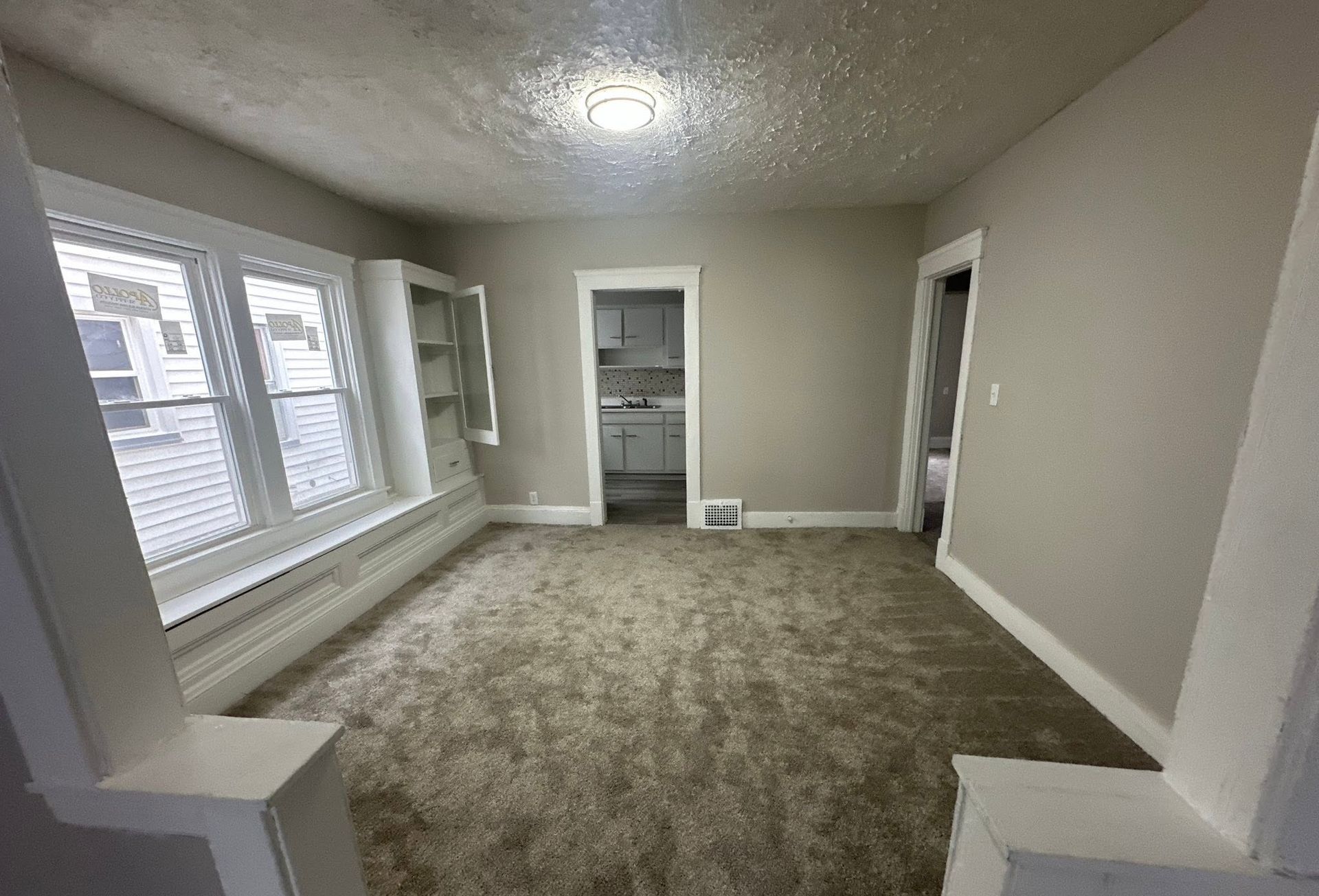 Empty beige living room with carpet, large windows, and a built-in alcove.