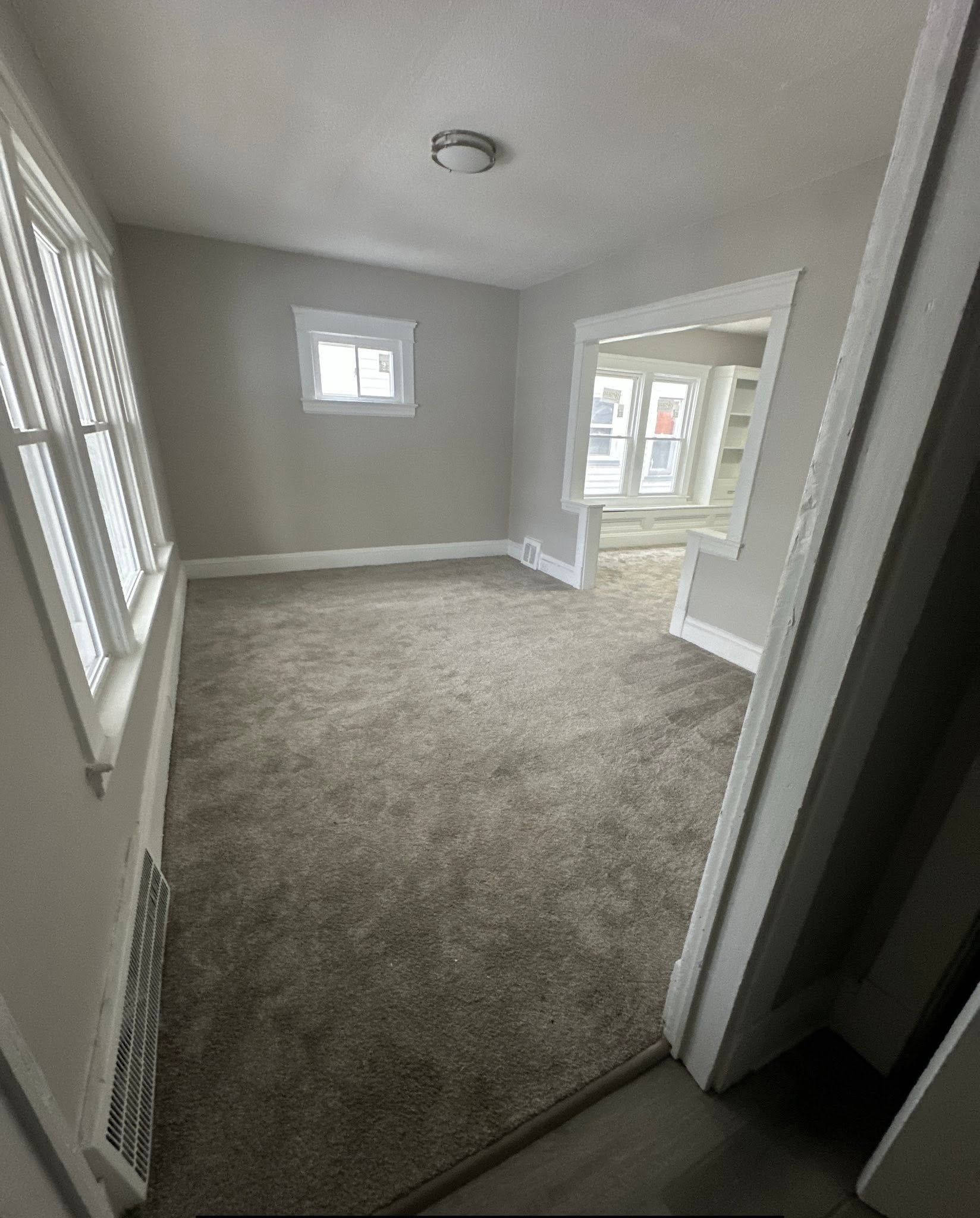 Empty gray carpeted room with white trim, small window, and doorway to a bright adjoining space