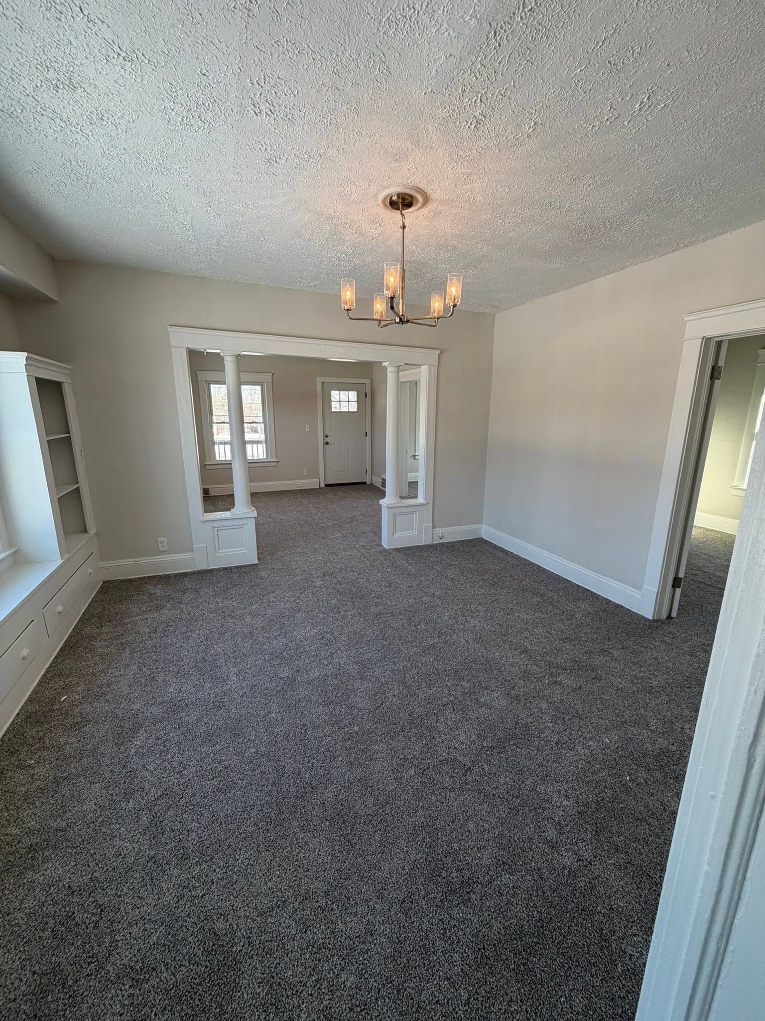 Empty carpeted living room with light gray walls, ceiling light, and doorway to another room