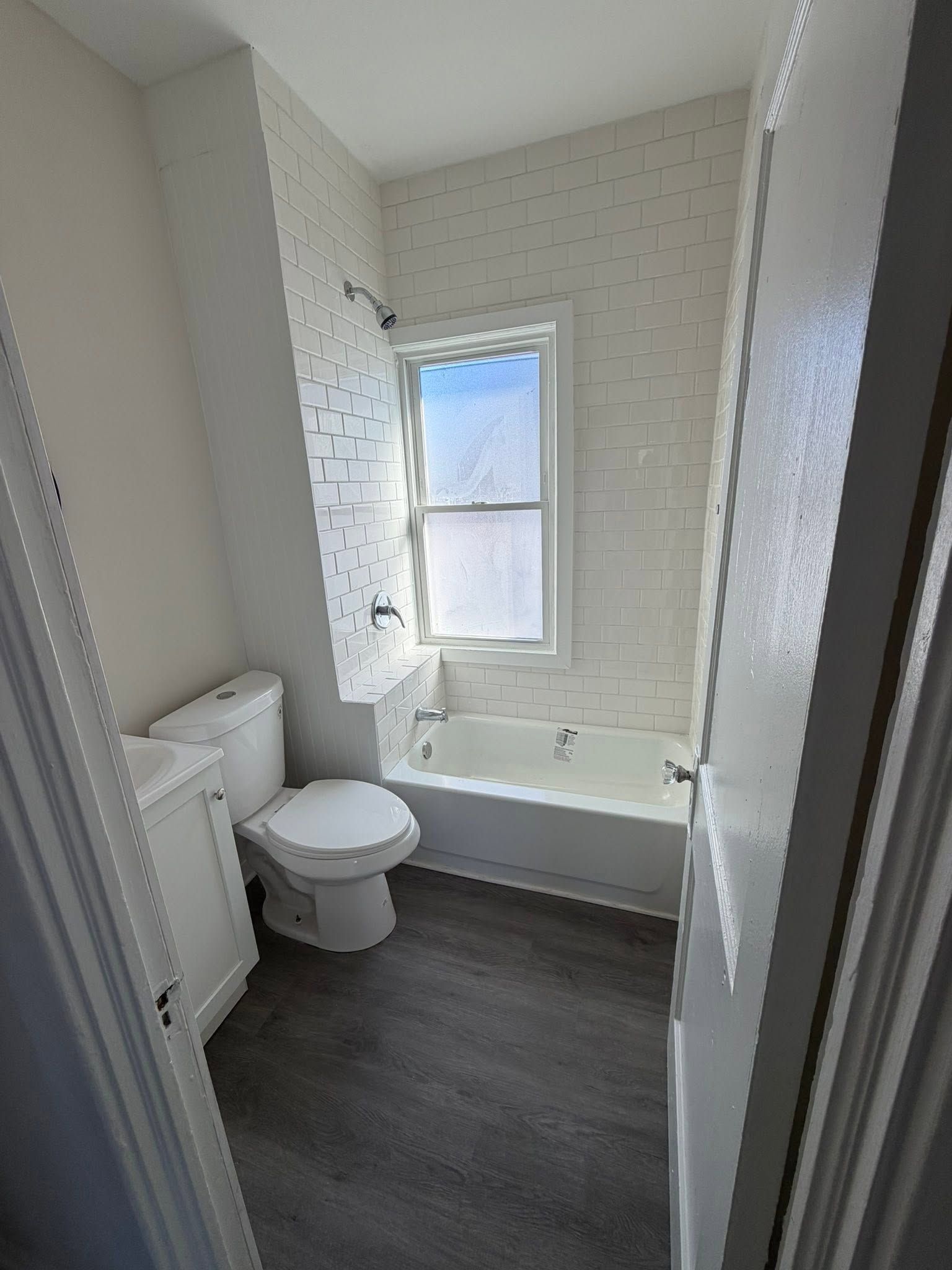 Small white bathroom with toilet, radiator, tub, window, and dark floor.