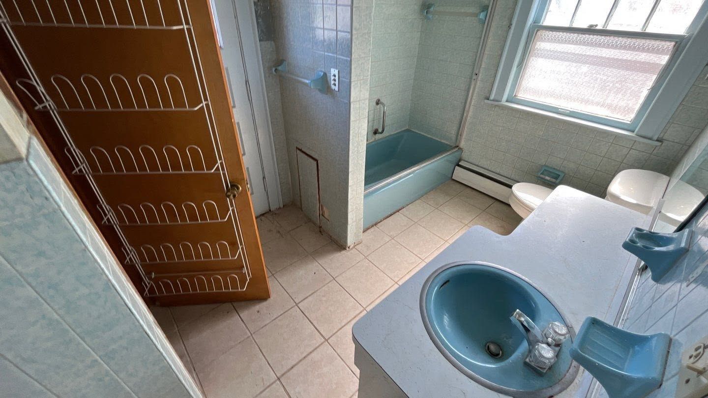 Small tiled bathroom with blue sink, toilet, window, and wooden cabinet.