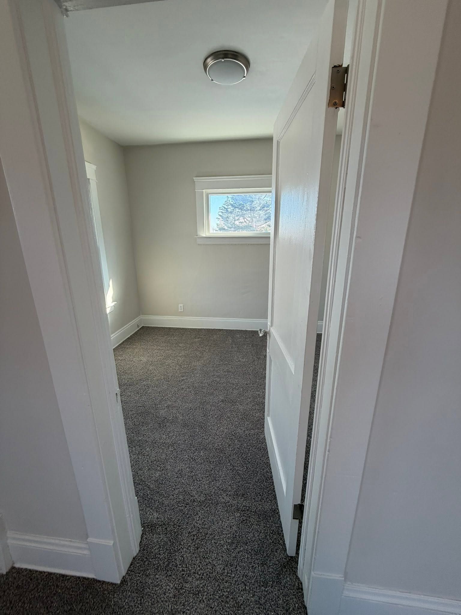 Narrow hallway with gray carpet, white walls, and a small window at the far end
