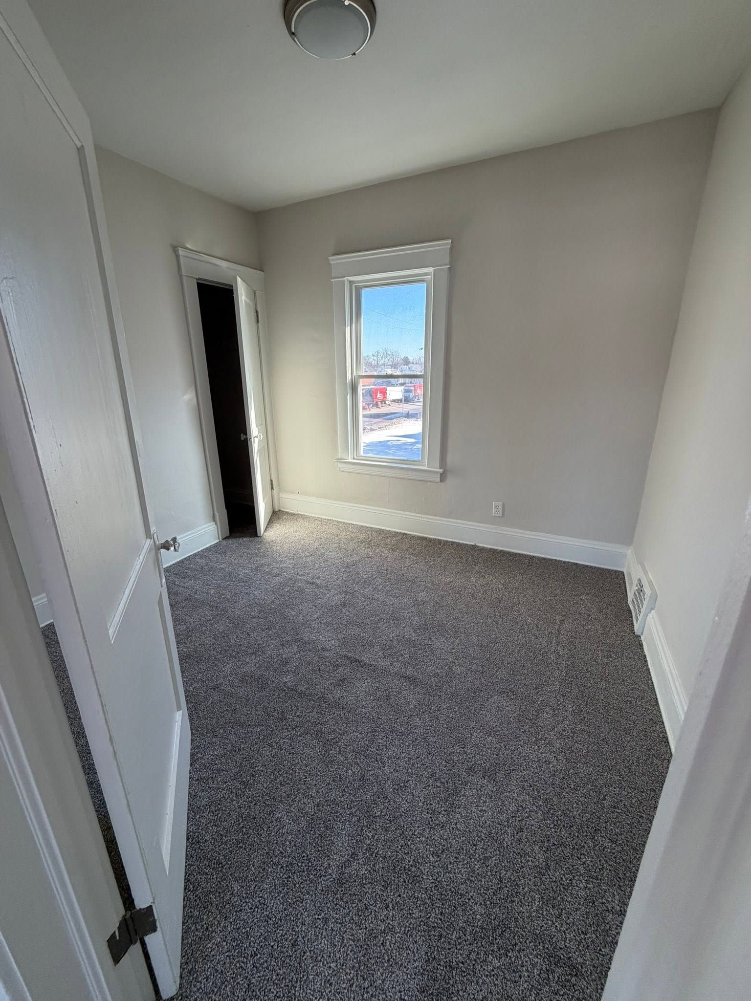 Empty carpeted room with beige walls, white trim, and a small window letting in daylight.