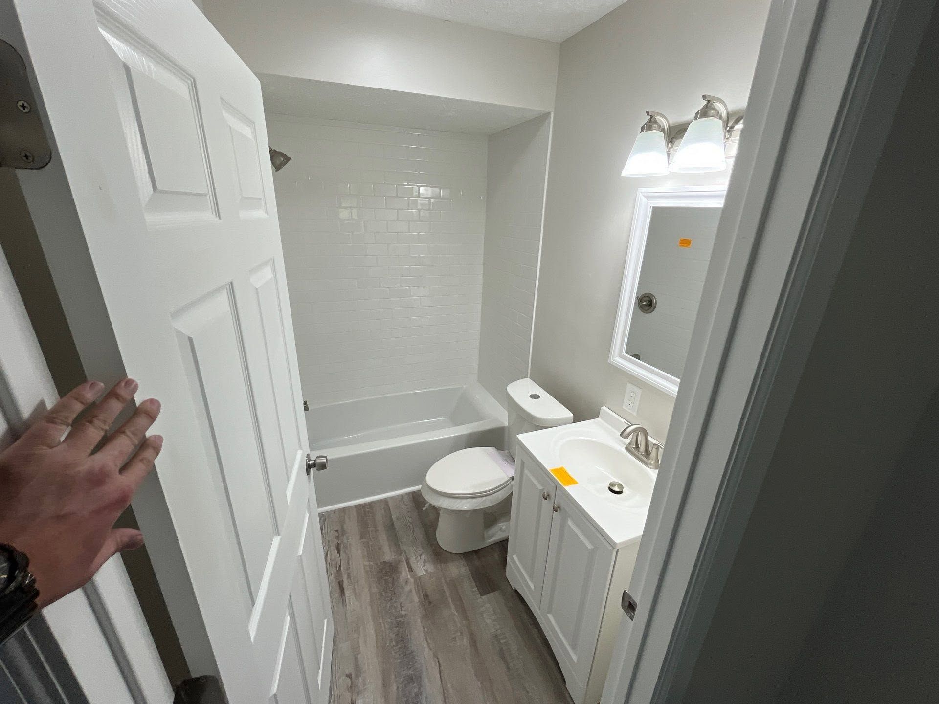 Small bathroom with white vanity, toilet, tub, and gray wood-look floor; hand holds the door open.