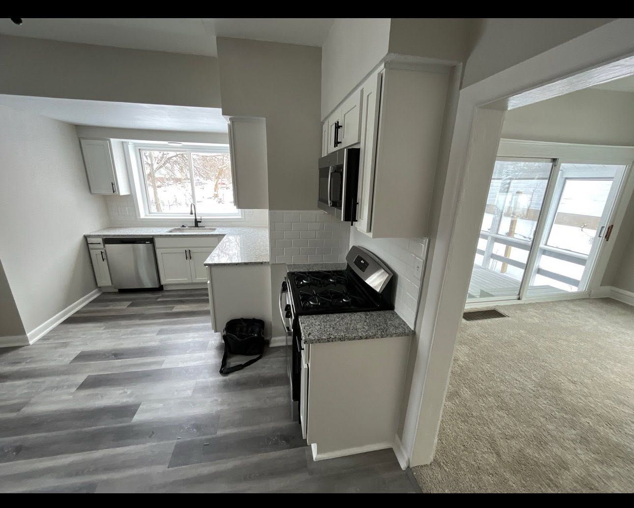 Bright kitchen with gray flooring, white cabinets, and a stove beside an open doorway to a carpeted room.