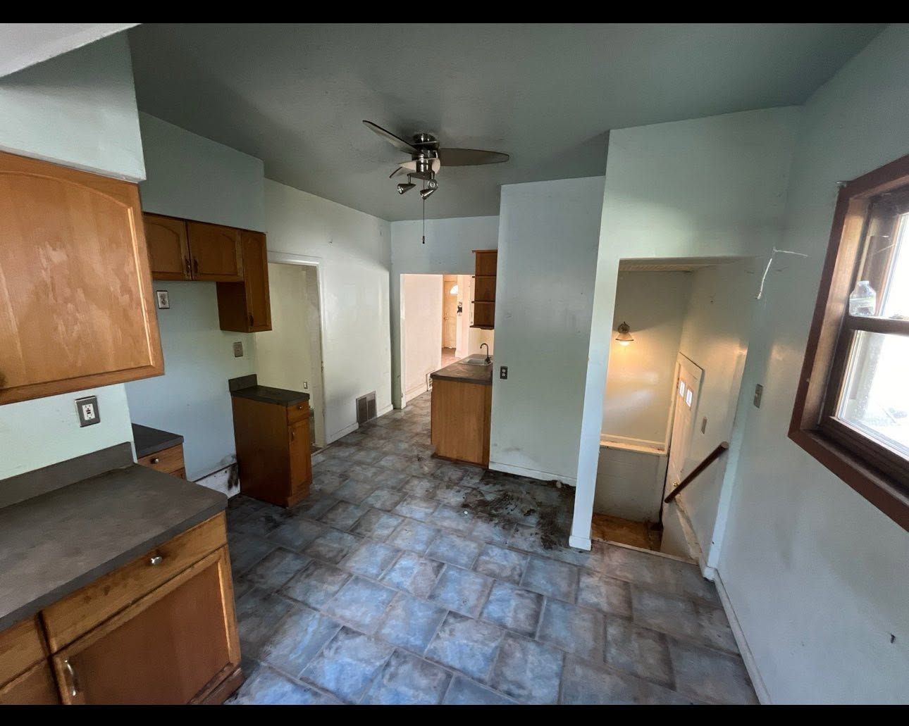 Empty kitchen with blue tile floor, wooden cabinets, ceiling fan, and stairs by a window.