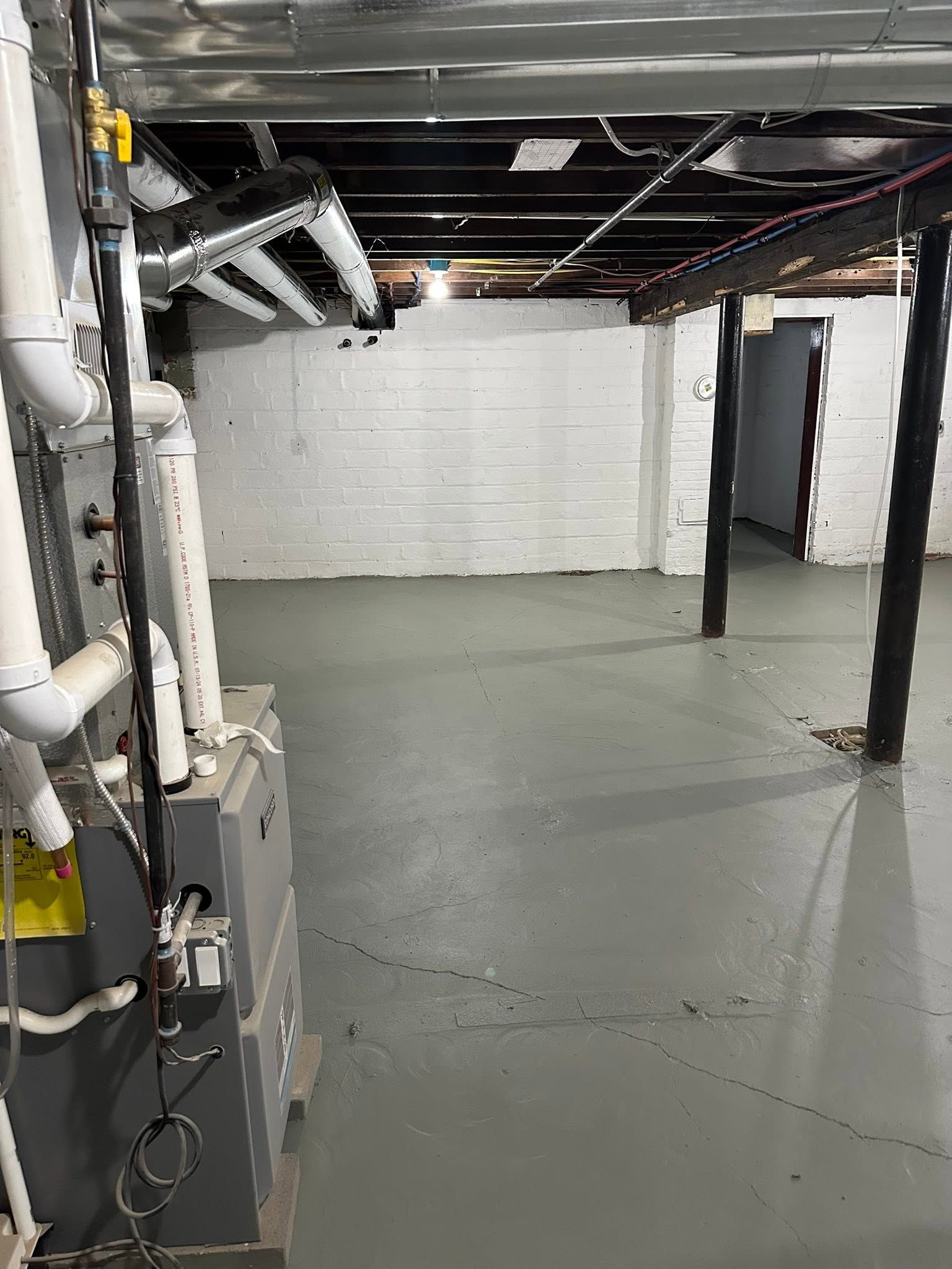 Empty basement with white brick walls, gray floor, exposed pipes, and support columns.