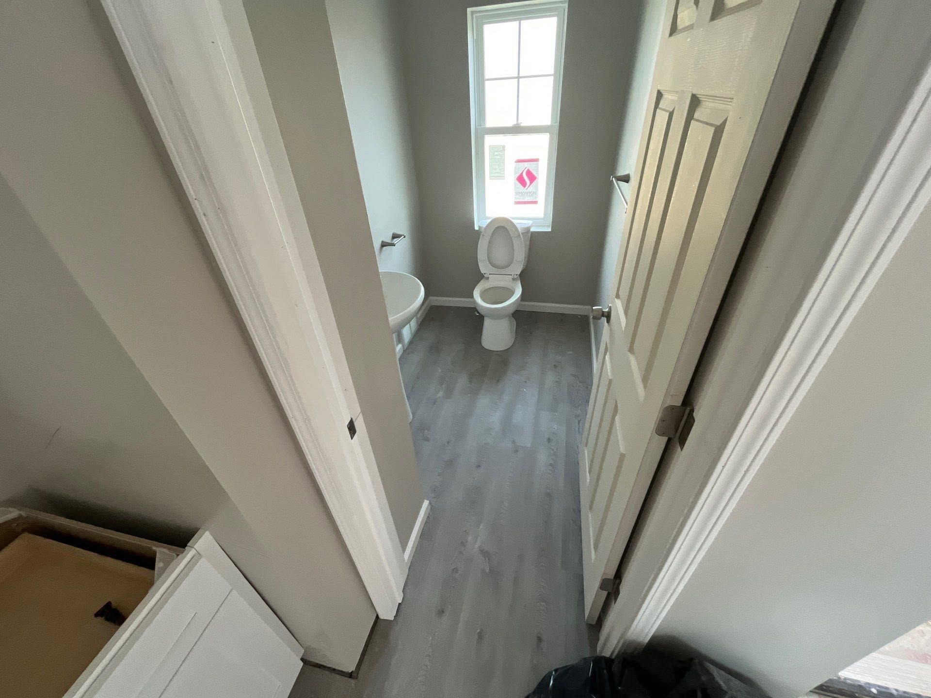 Narrow hallway leading to a small bathroom with white walls, gray floor, and a toilet by a window.