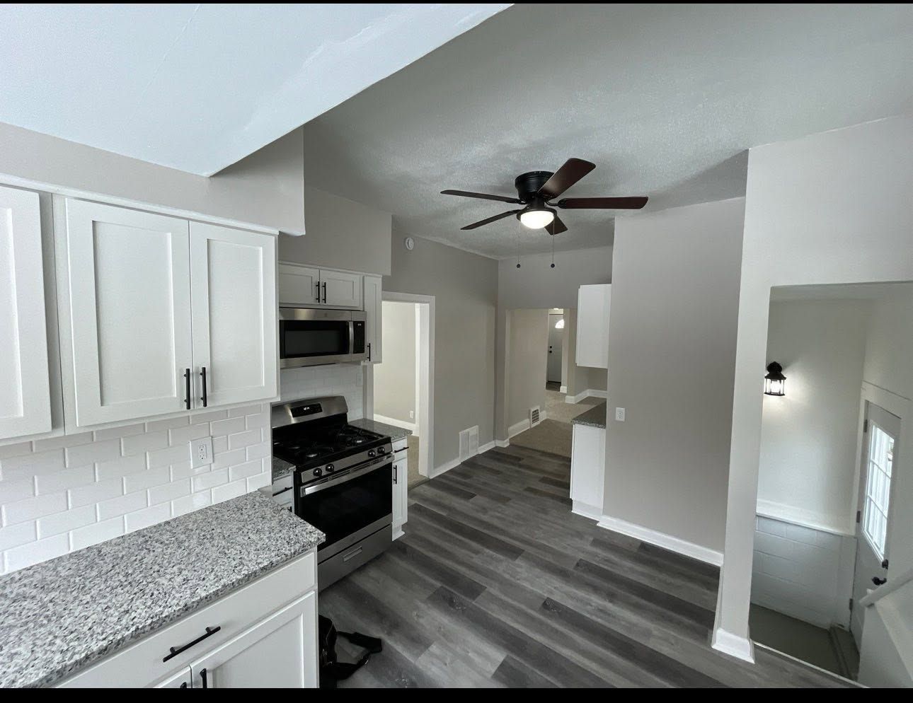 Modern white kitchen with gray countertops, stainless stove, gray plank flooring, and ceiling fan leading to a hallway