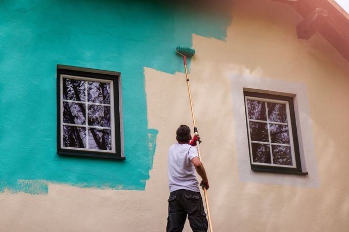 Person painting a wall with teal and peach sections, using a roller near two windows.