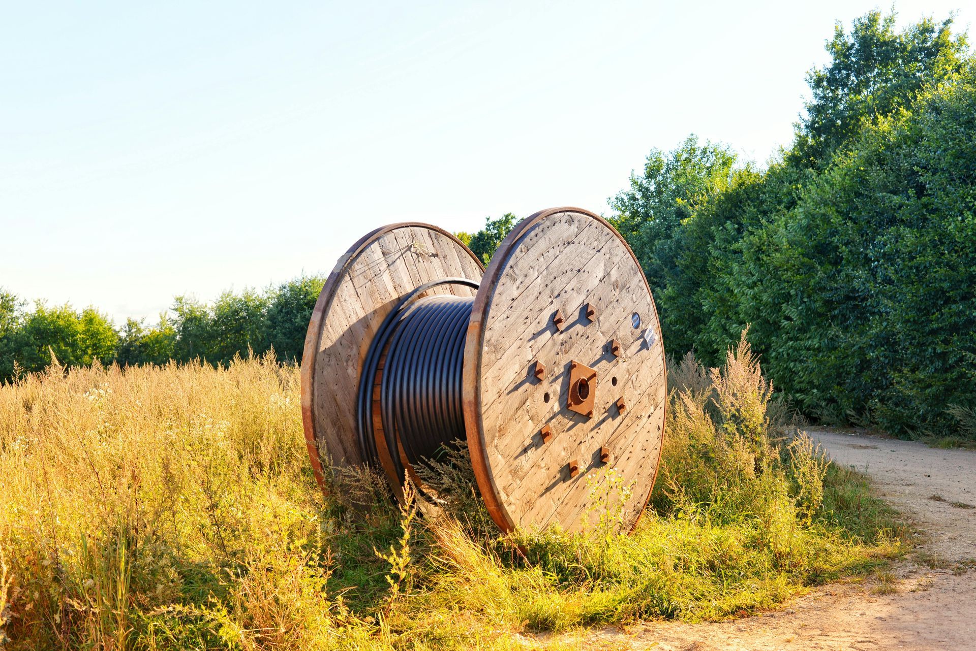 Grote houten kabelhaspels naast een zandpad in een zonnig grasveld, met bomen op de achtergrond.