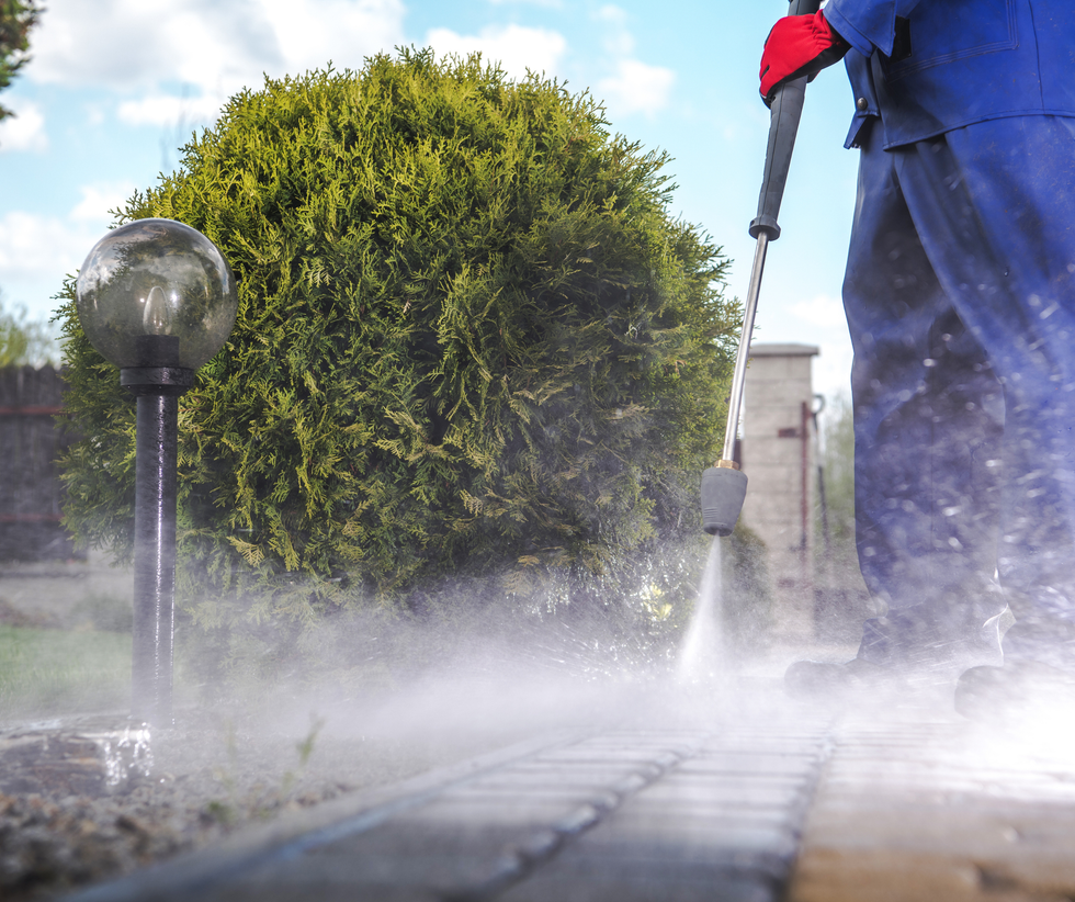 Person pressure-washing a patio, with mist spraying beside a trimmed bush and lamp post
