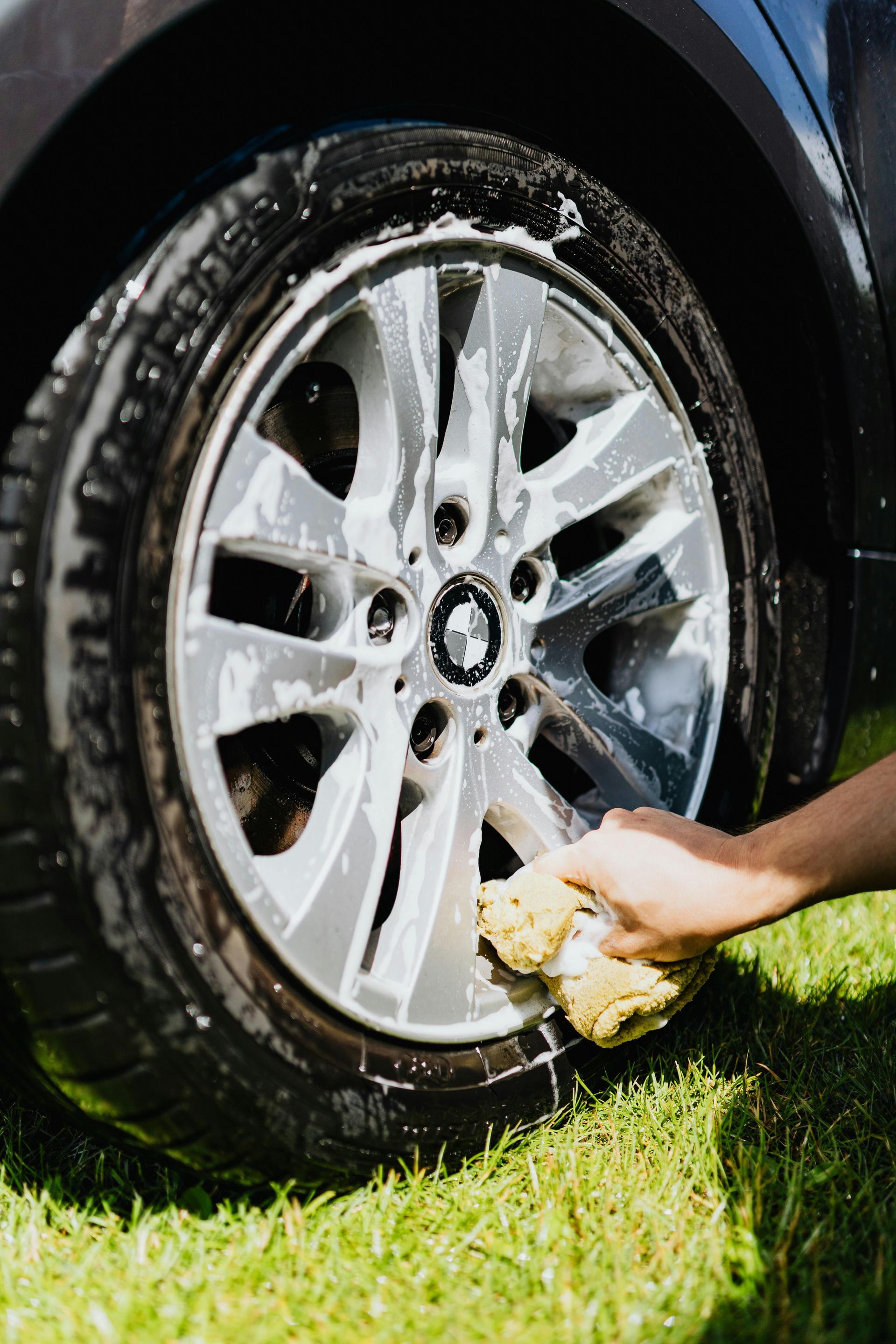 Hand washing a car wheel with a sponge on a grassy lawn.