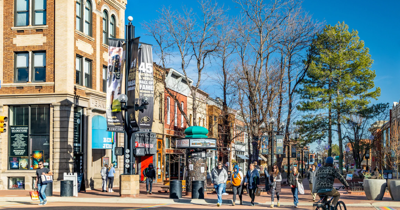 Street scene with shops, pedestrians, trees, and a bus shelter on a sunny day.
