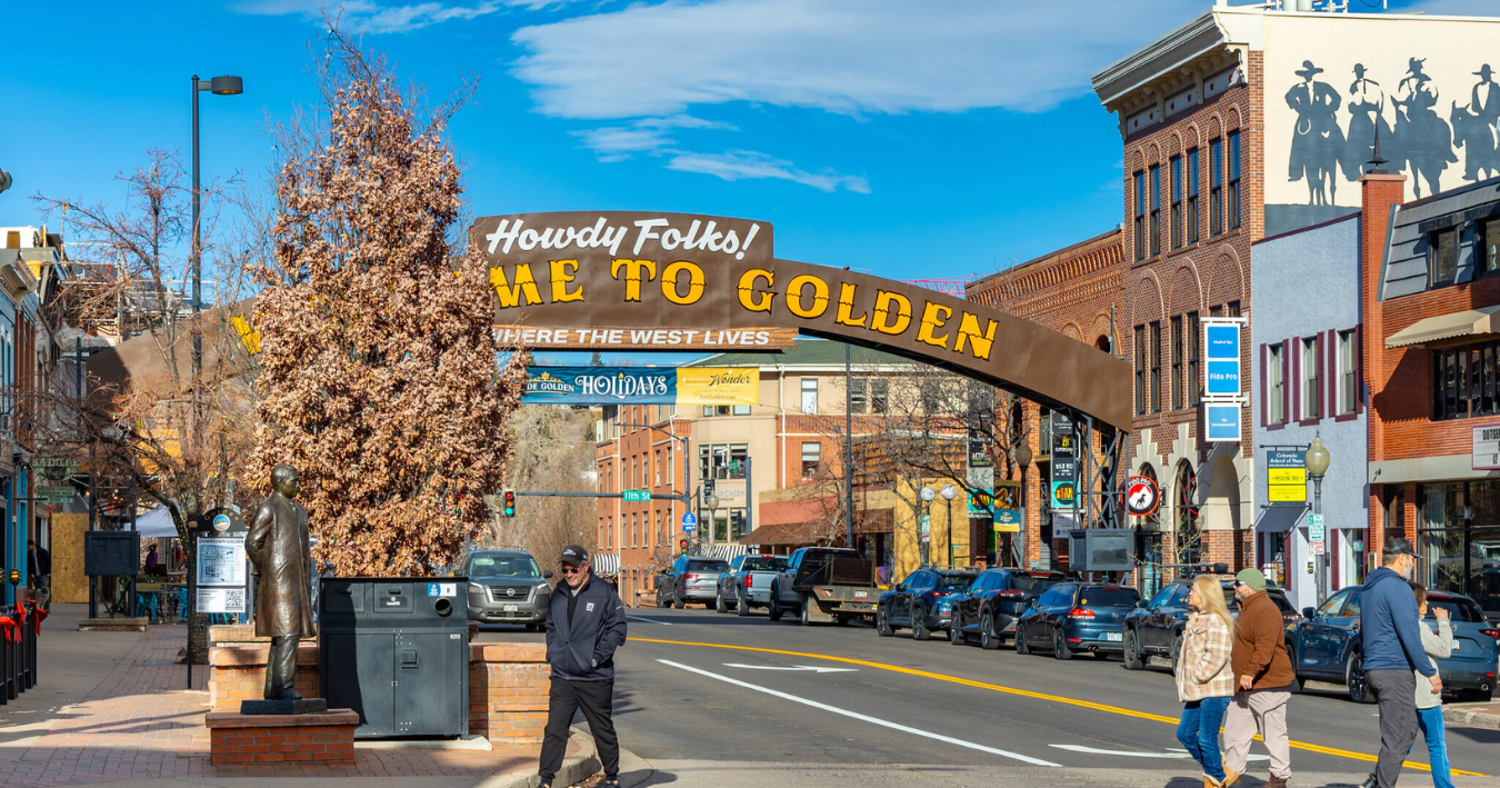 Archway welcoming visitors to Golden, Colorado, with buildings and parked cars. People walking on a crosswalk.
