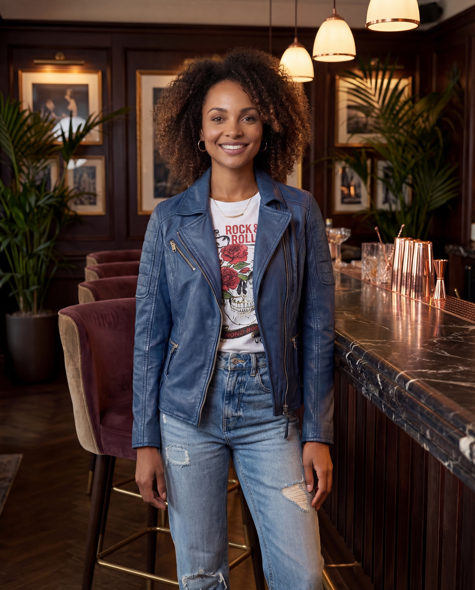 Person standing in a stylish lounge, smiling in a denim jacket and jeans beside a marble bar.