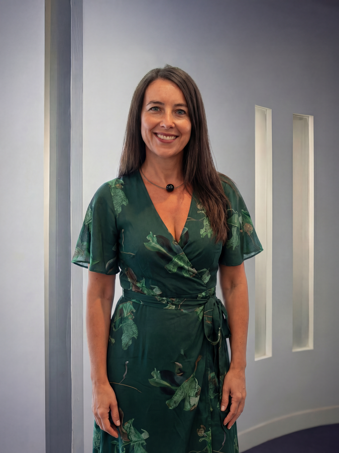 Woman in green floral wrap dress smiles, standing indoors near a light-colored wall. Account & Style Director at ACi Studios.