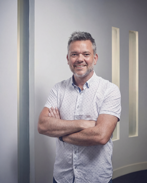 Giles Mosley standing with arms crossed in a bright hallway, wearing a white patterned shirt and smiling.