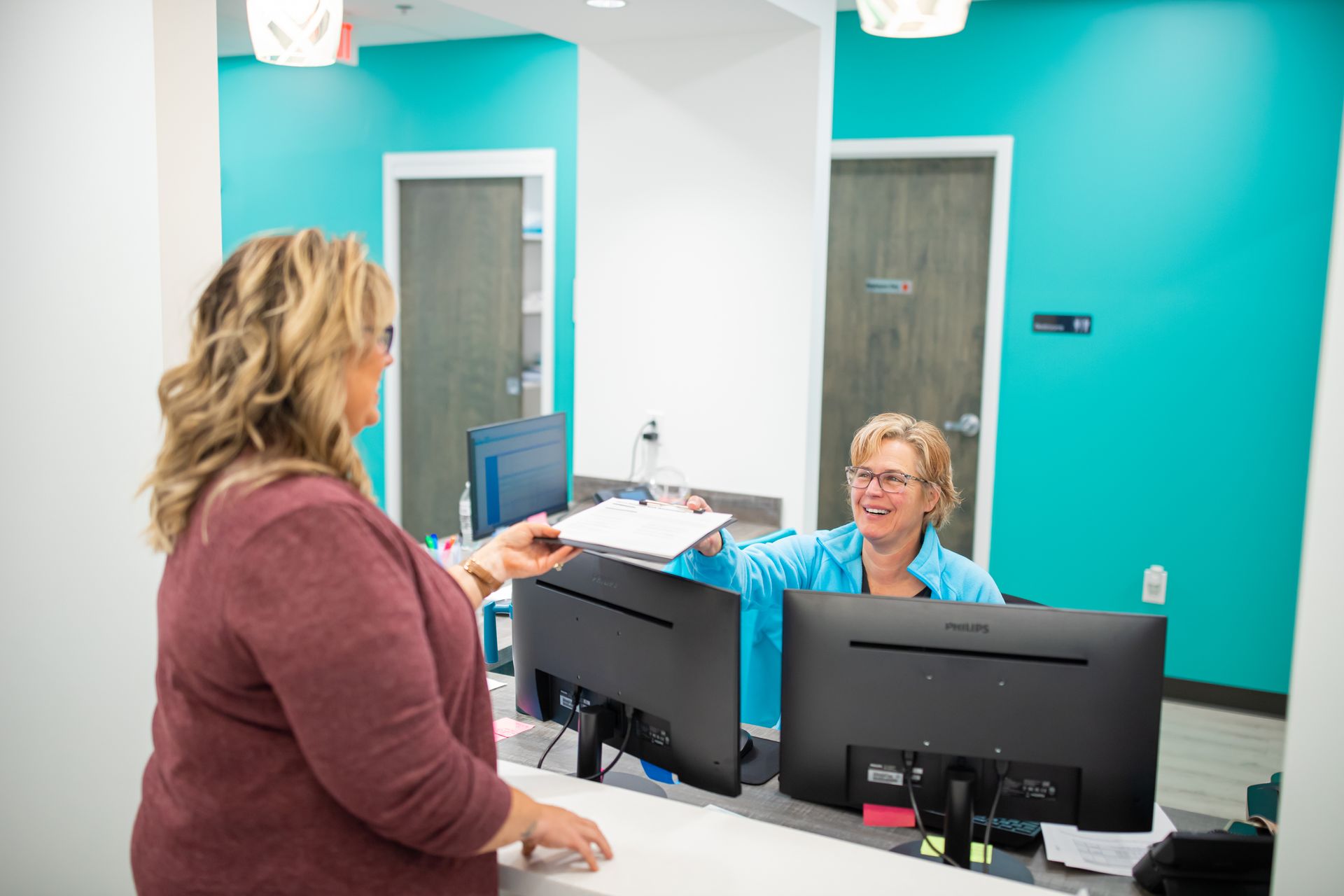 A woman hands paperwork to a receptionist behind a teal desk in an office setting.
