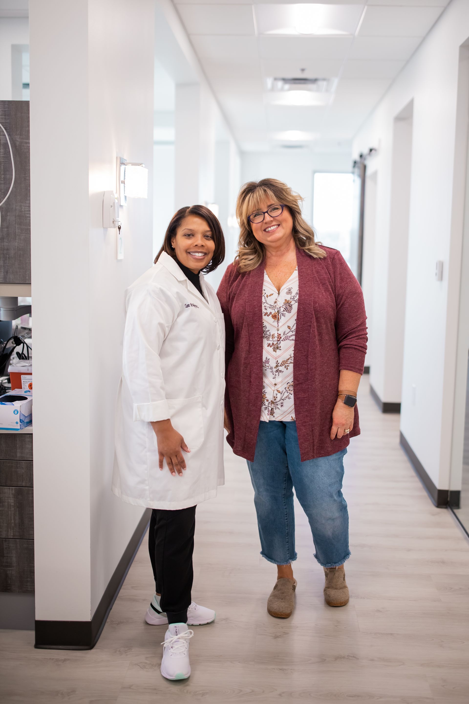 Two women smiling in a well-lit hallway. One in a lab coat, the other in a cardigan and jeans.
